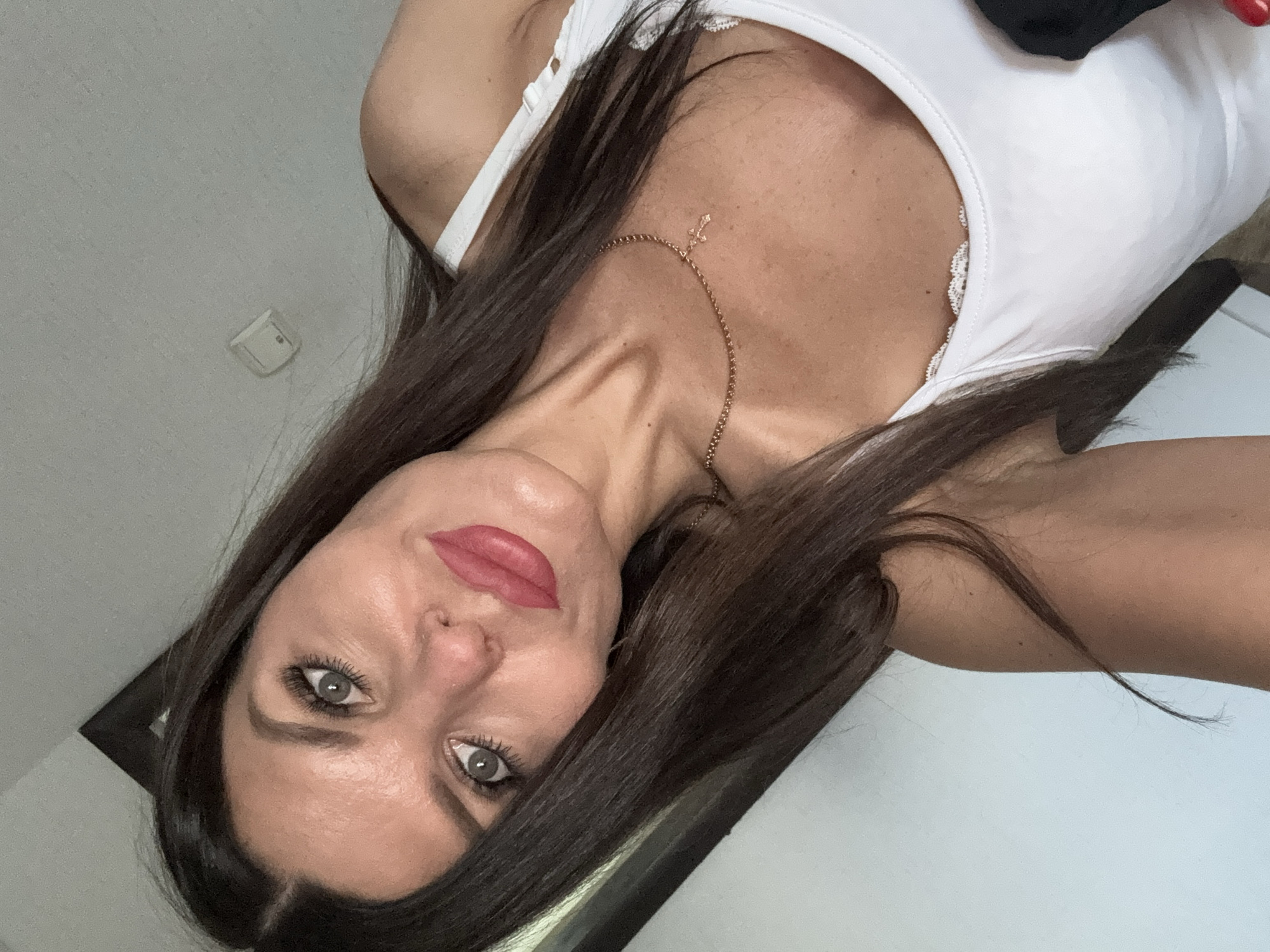 A woman with long, dark brown hair and grey eyes is posing for a selfie indoors. She is wearing a white tank top with lace trim and a delicate gold necklace with a small cross pendant. Her lips are painted a vibrant pink. The background appears to be a room, with a wall and what looks like a doorframe visible. There is no indication of the city of Chaikovsky, Russia, in this image.