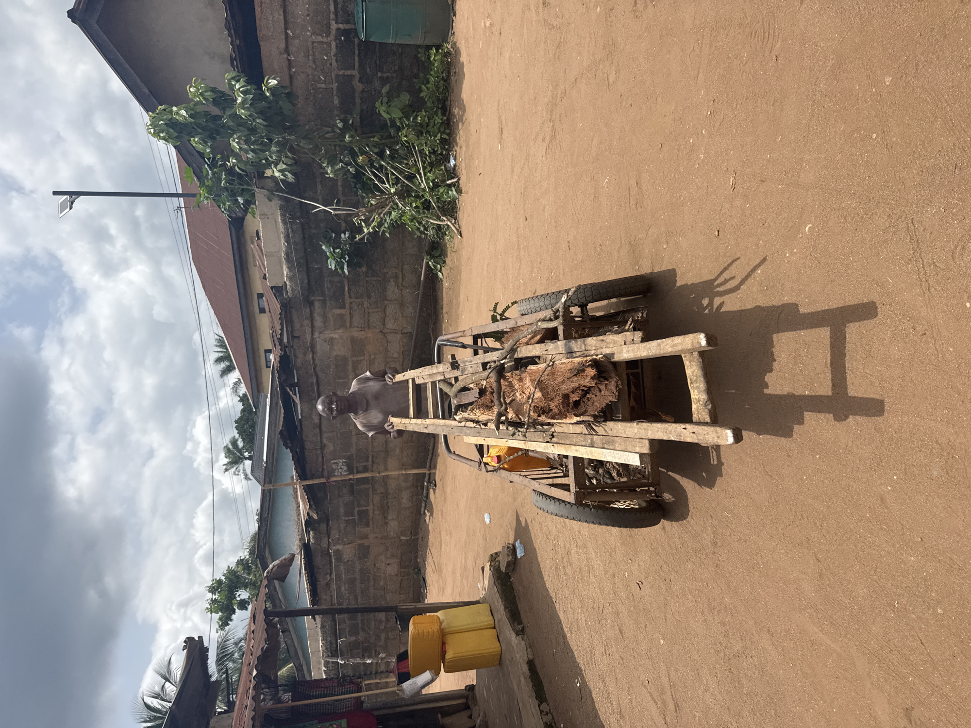 The image depicts a man in Aboh, Nigeria, pulling a cart laden with what appears to be palm fronds or a similar fibrous material, down a dirt road. The surroundings include buildings with weathered walls, corrugated metal roofs, vegetation, and stacked yellow jerrycans. The sky is overcast.