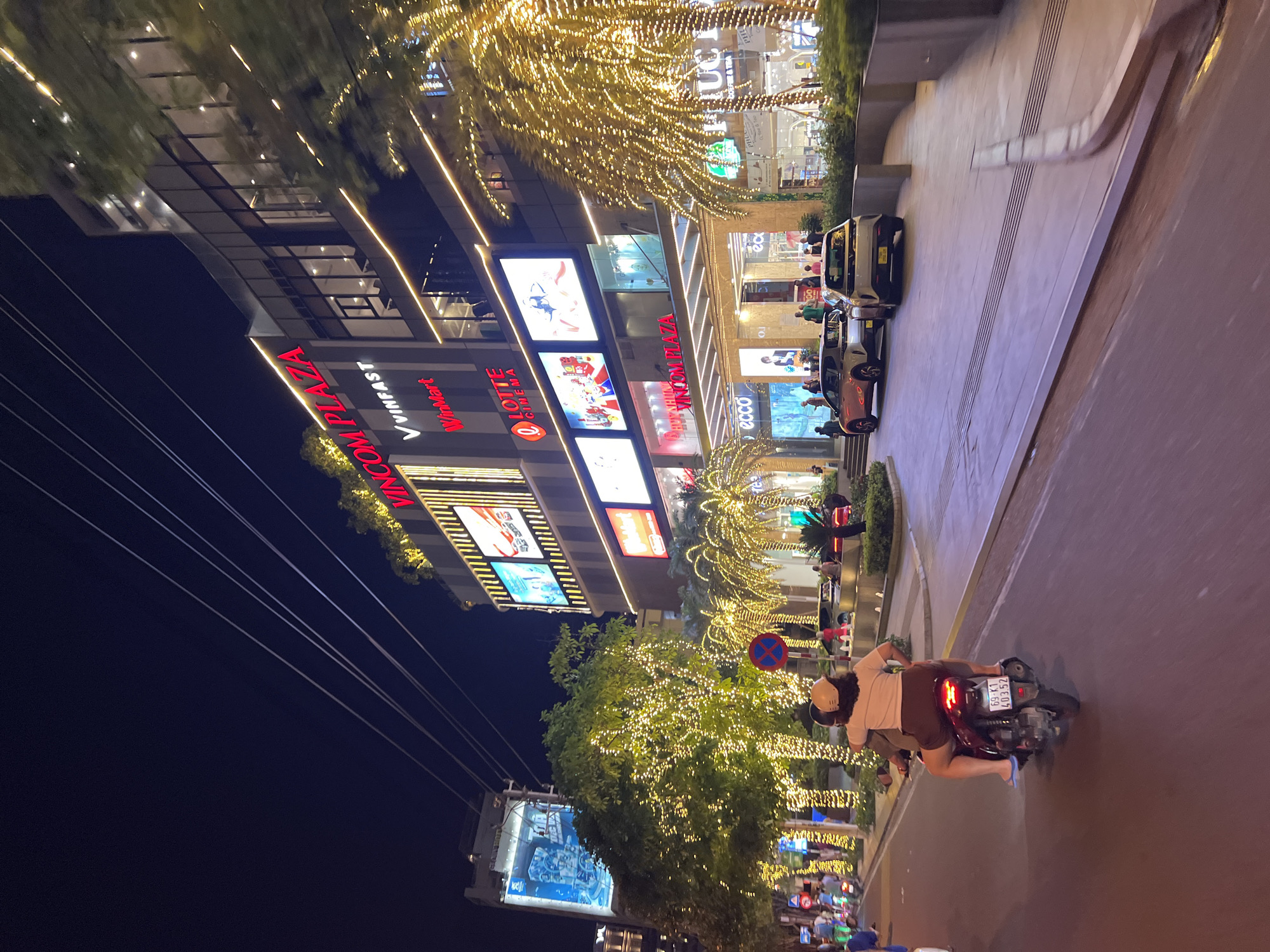 The scene is an urban street at night, likely in Nha Trang, Vietnam, given the presence of "VINCOM PLAZA". A multistory building is illuminated with lights and signs for "Vinfast," "Winmart," and "Lotte Cinema." Palm trees decorated with lights line the building and street. A motorcycle is driving on the right side of the street. The rider is wearing a helmet and a light-colored shirt and brown shorts. A passenger is also riding on the motorcycle. The motorcycle's license plate reads "69-K1 403.52." Pedestrians and other streetlights are visible further down the street.