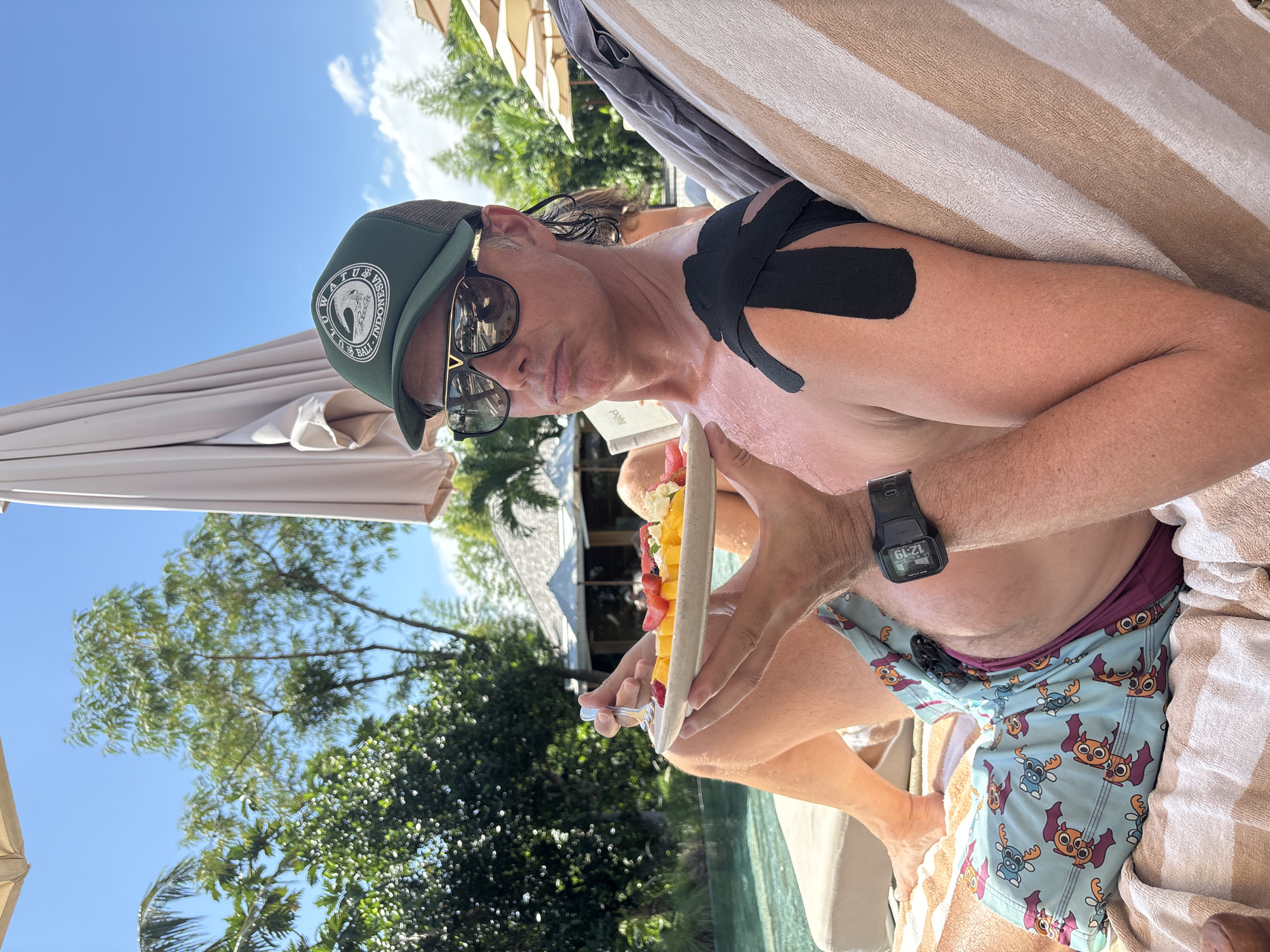A man is sitting outdoors, possibly on a lounge chair, eating fruit from a long, narrow plate. He's wearing a green baseball cap with a circular logo on the front, sunglasses, and a black digital watch on his left wrist. Black kinesiology tape is applied to his left shoulder. He's wearing light blue swim trunks with a cartoon character pattern. He's holding a fork in his right hand. The background includes trees, an umbrella, and a building, suggesting a tropical resort or vacation setting. The hat logo mentions "WATU VISSENESSA INDONESIA BALI," indicating the location is likely near or within Bali, Indonesia. North Kuta, Indonesia is the identified city.
