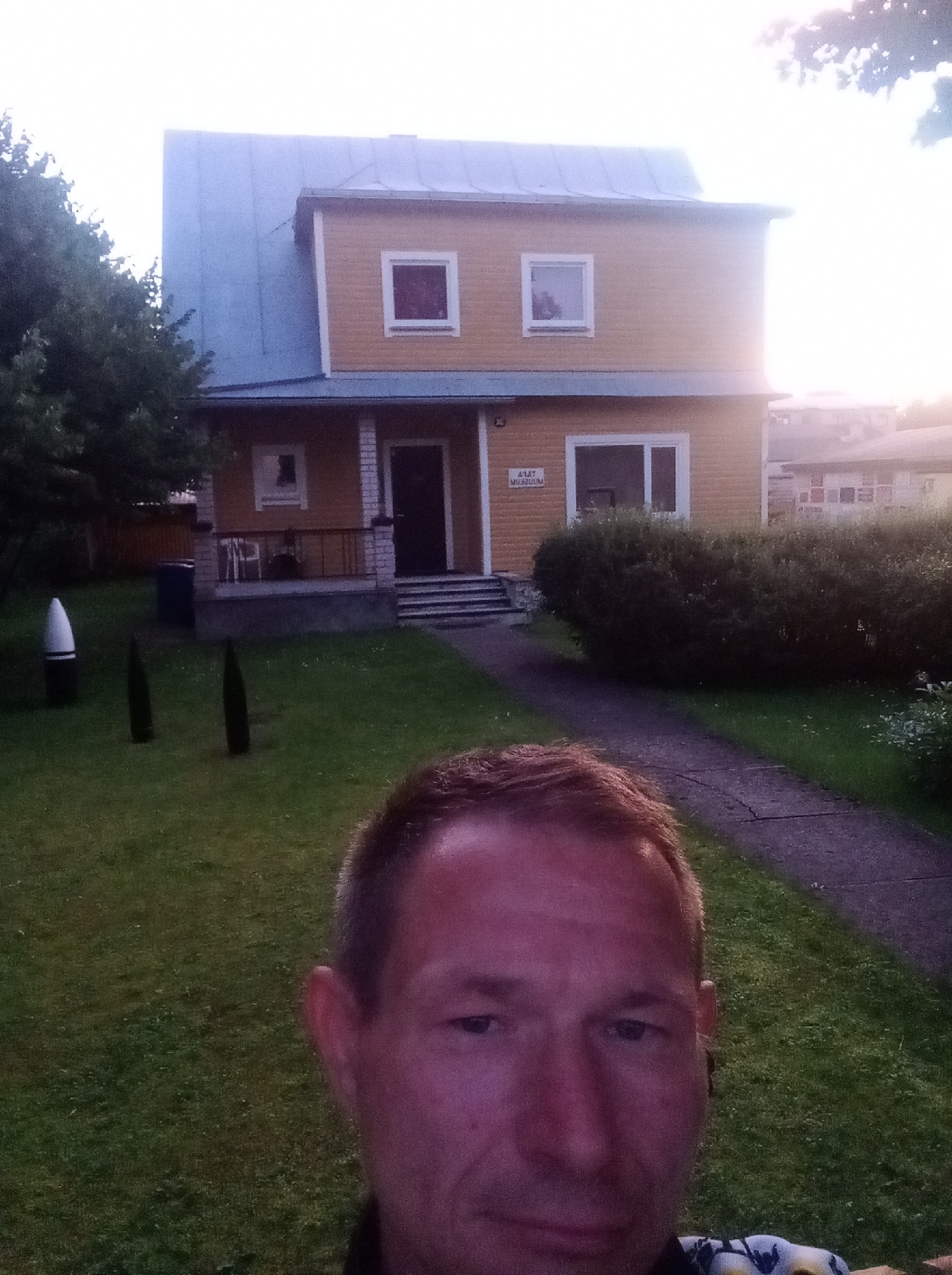 In Tapa, Estonia, a man takes a selfie in front of a two-story yellow house with a light blue metal roof. The house has white-trimmed windows and a dark-colored door accessible by a set of concrete steps. A sign above the ground-floor window reads "AJAT MUUSEUM." To the left of the house, in the grassy yard, are three dark, conical objects and a white and black cylindrical object. A paved walkway leads from the house's entrance through the lawn. The scene is illuminated by the soft light of what appears to be late afternoon or early evening sun.
