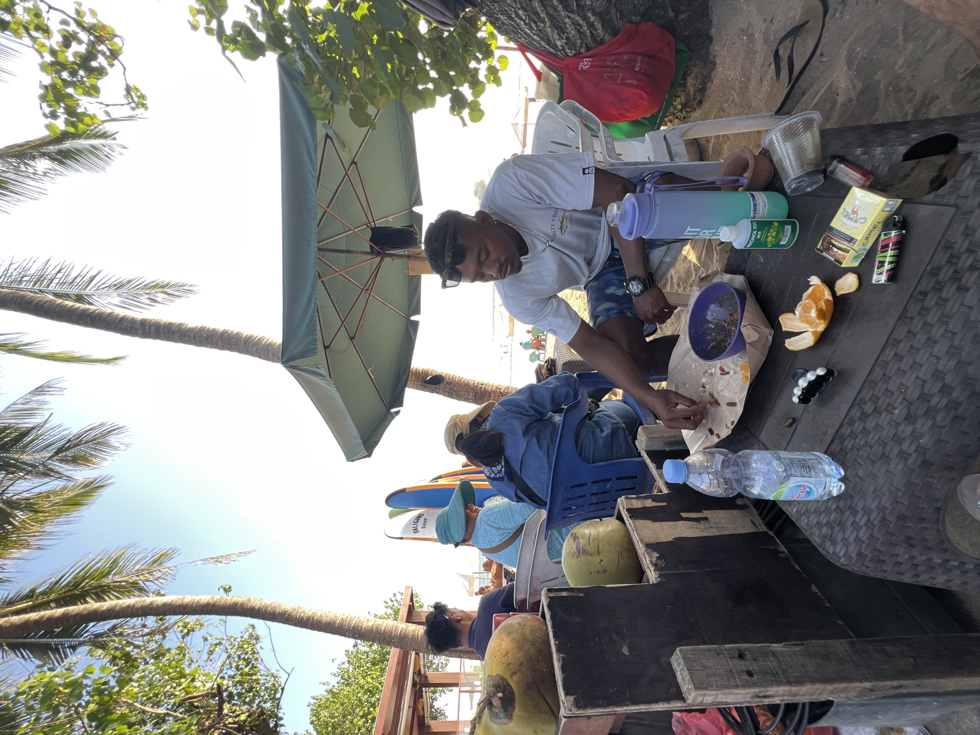 An outdoor scene on a beach in Kuta, Indonesia, features several people gathered around a table. A man wearing sunglasses and a grey t-shirt sits on a white plastic chair under a green umbrella, appearing to eat. In front of him on a wicker table are a purple bowl with leftover food, an orange peel, a pack of Camel cigarettes, a lighter, a bottle of hand sanitizer, and a tall purple and blue water bottle. A clear plastic bottle of water is also visible. Behind him, several other individuals are seated, some wearing hats. Two coconuts are placed on a wooden structure to the left. Surfboards are visible in the background. The ground is sandy, consistent with a beach environment. The lighting suggests it is daytime, likely late afternoon or early morning due to the low sun angle.