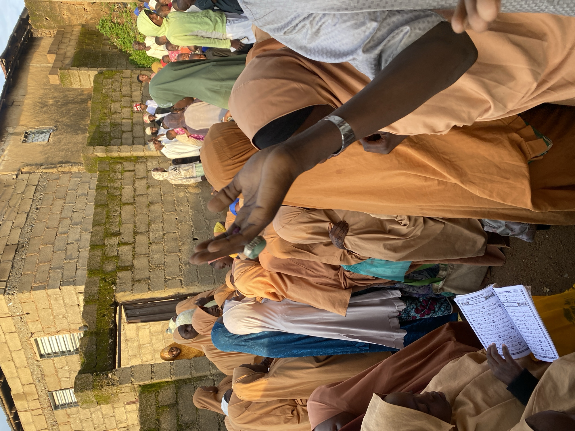 In Jos, Nigeria, a group of people, primarily children, are gathered in an outdoor courtyard. They are dressed in traditional Islamic attire, with many wearing colorful hijabs and robes in shades of orange, tan, and blue. Some individuals are reading from books, possibly religious texts, while others appear to be listening or observing. The setting is rustic, with buildings constructed from concrete blocks and a dirt ground. The overall atmosphere suggests a community gathering or educational event.