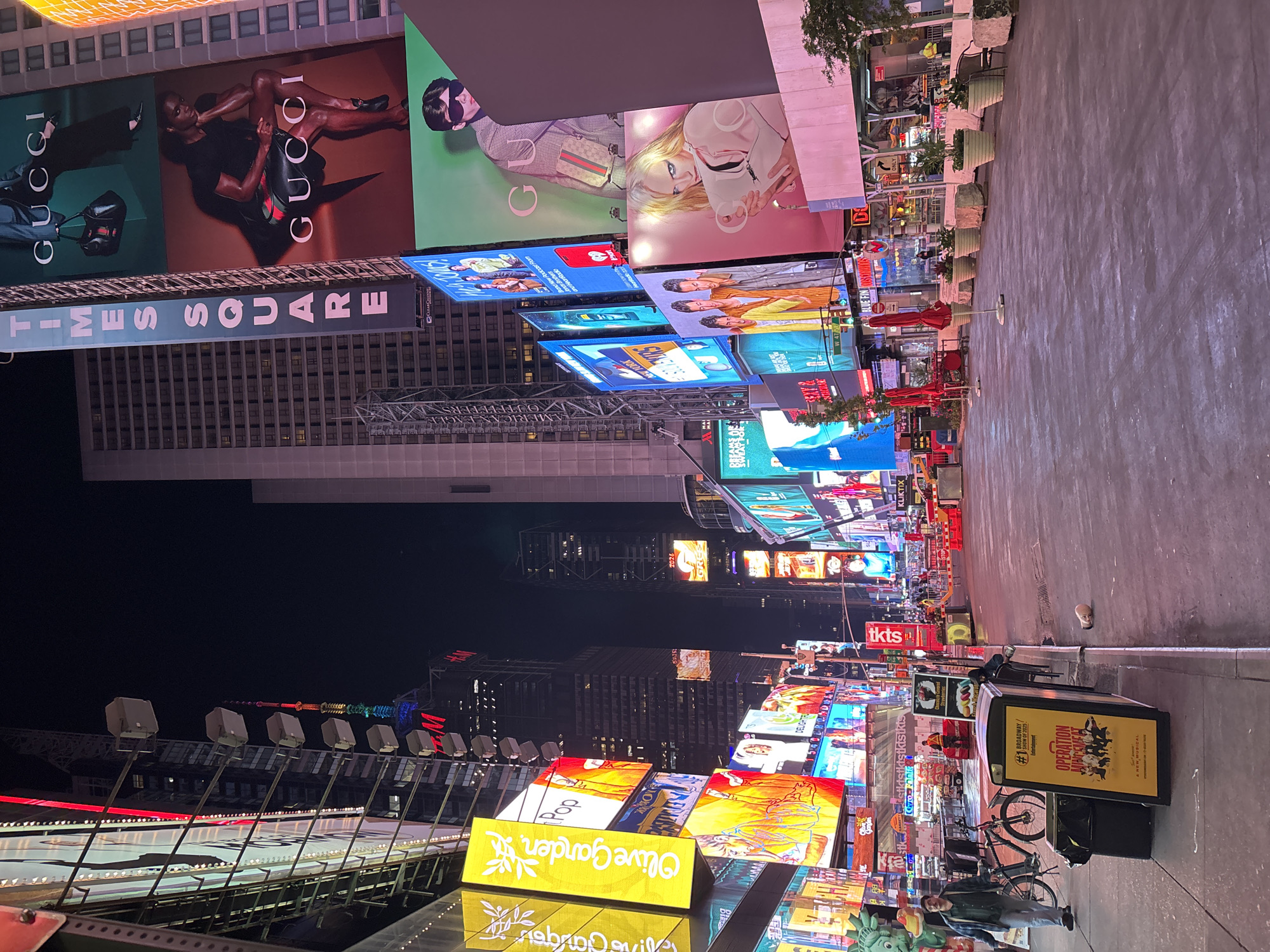 This photograph captures a vibrant nighttime scene in Times Square, New York City, dominated by a multitude of brightly lit digital billboards and static advertisements. The image is taken from a slightly elevated perspective, looking down a wide pedestrianized street lined with buildings that are themselves adorned with massive displays.
The primary subjects are the illuminated advertisements, which showcase various brands and products. Prominently featured are large "GUCCI" billboards displaying models, one showing a man in a black shirt and shorts, and another a person in a patterned jacket with a handbag. A "TIMES SQUARE" sign is visible below the Gucci ads. Further down the street, an "Olive Garden" sign stands out with its yellow background, and other screens display vibrant graphics and text such as "Pop" and "DELIGHTFUL." In the mid-ground, a "tkts" booth is identifiable, and nearby, a yellow kiosk advertises "OPERATION NEW MUSICAL" for "Entertainment." A bicycle is parked beside this kiosk, and a person can be seen standing nearby.
The setting is an iconic urban environment, characteristic of Times Square, with towering buildings forming the backdrop. The time of day is clearly night, as indicated by the dark sky, making the artificial lights of the billboards, streetlights, and building illuminations the dominant light sources. The street appears clear, suggesting fair weather. Planters with greenery line parts of the walkway, and red umbrellas from what might be an outdoor seating area add splashes of color.
Other visible text includes "2025," "2026," "KLIKTIX," "@stkstkistkistktstkts," "Gyro-Kebab," "Brand New Podcast," "NEW YORK," and partially visible names like "American Eagle Outfitters" and "H&M." The overall atmosphere is one of bustling commercial energy and overwhelming visual stimulation.
