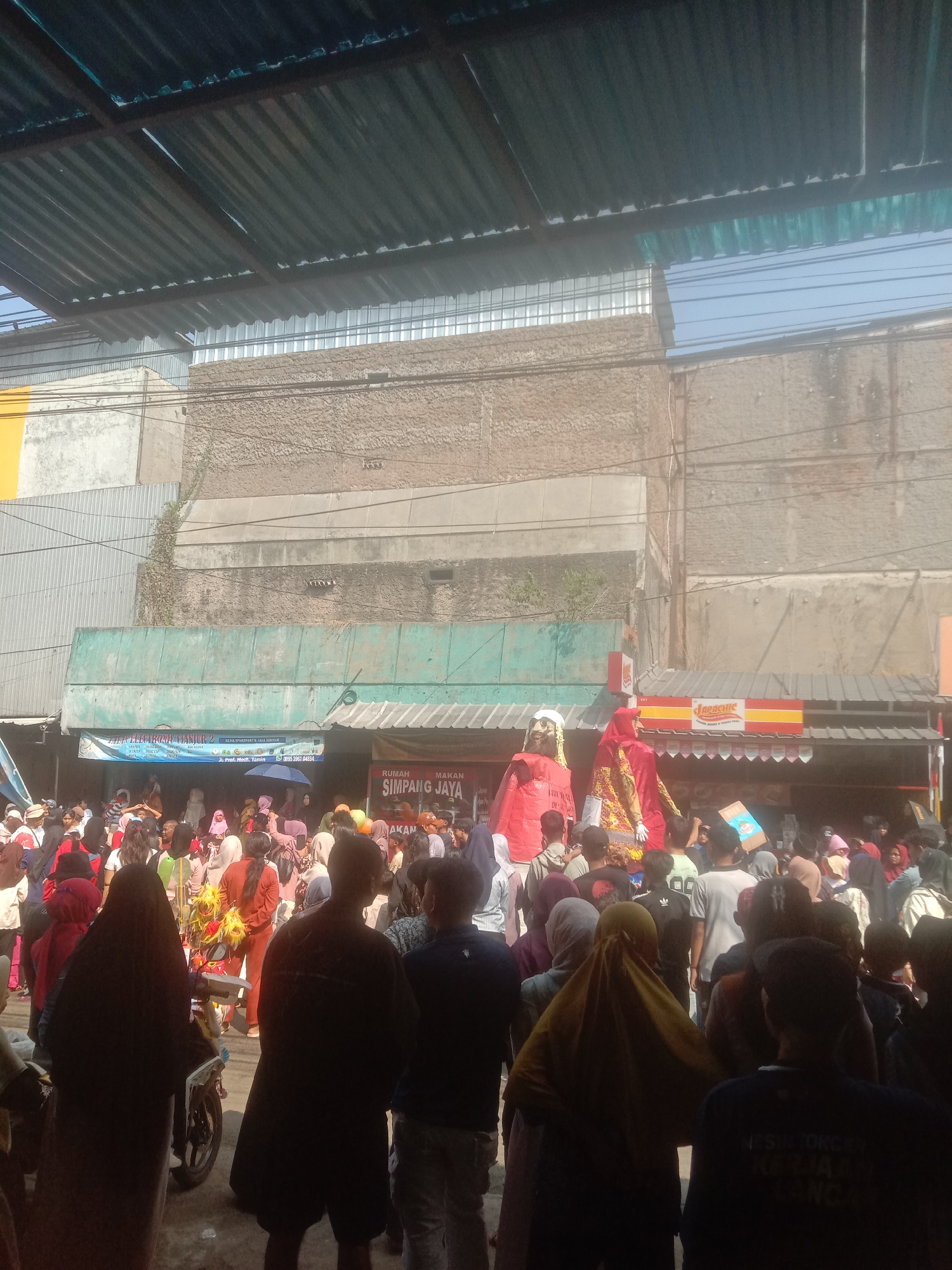 The image depicts a crowded street scene in Cianjur, Indonesia, likely during a local festival or event. Two large, elaborately dressed figures, resembling giants or puppets, are prominently displayed at the center of the gathering, suggesting a cultural performance or parade. A dense crowd of people, many wearing traditional Indonesian attire such as hijabs and sarongs, surrounds these figures, indicating active participation and observation.
In the background, a variety of buildings and structures are visible, including what appears to be a shopfront with signage that partially reads "SIMPANG JAYA" and "Japachic". The architecture suggests a typical urban Indonesian environment with corrugated metal roofing and brick or concrete walls. A motorcycle is parked to the left of the frame, amidst the crowd. The overall atmosphere seems festive and energetic, with people engaging with the central spectacle.