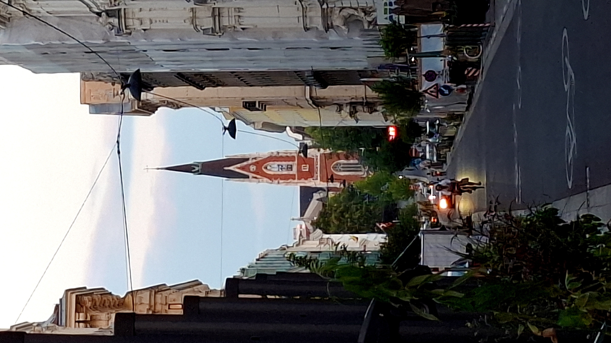 The image shows a street in Vienna, Austria, with a red church spire visible in the distance. The street is lined with buildings on both sides, and there is traffic and a person riding a bicycle. The lighting suggests it is either early morning or late evening.