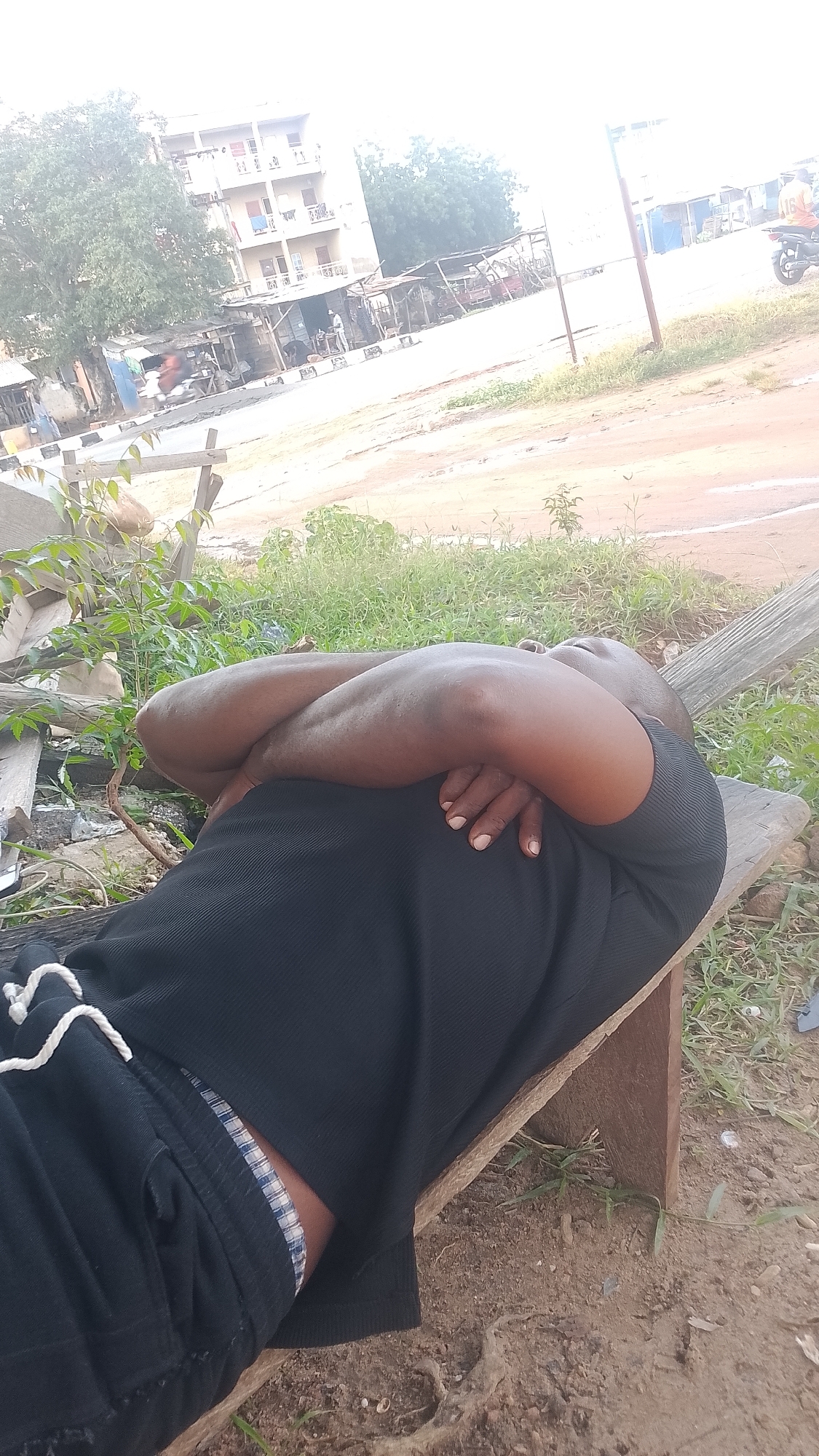 A man in black clothing is lying down on a wooden bench in Adankolo, Nigeria. His arms are crossed in front of him, and his head is resting on his arms. The man appears to be sleeping or resting. The background shows buildings, trees, and a dirt road. The scene suggests a warm, possibly dusty environment.
