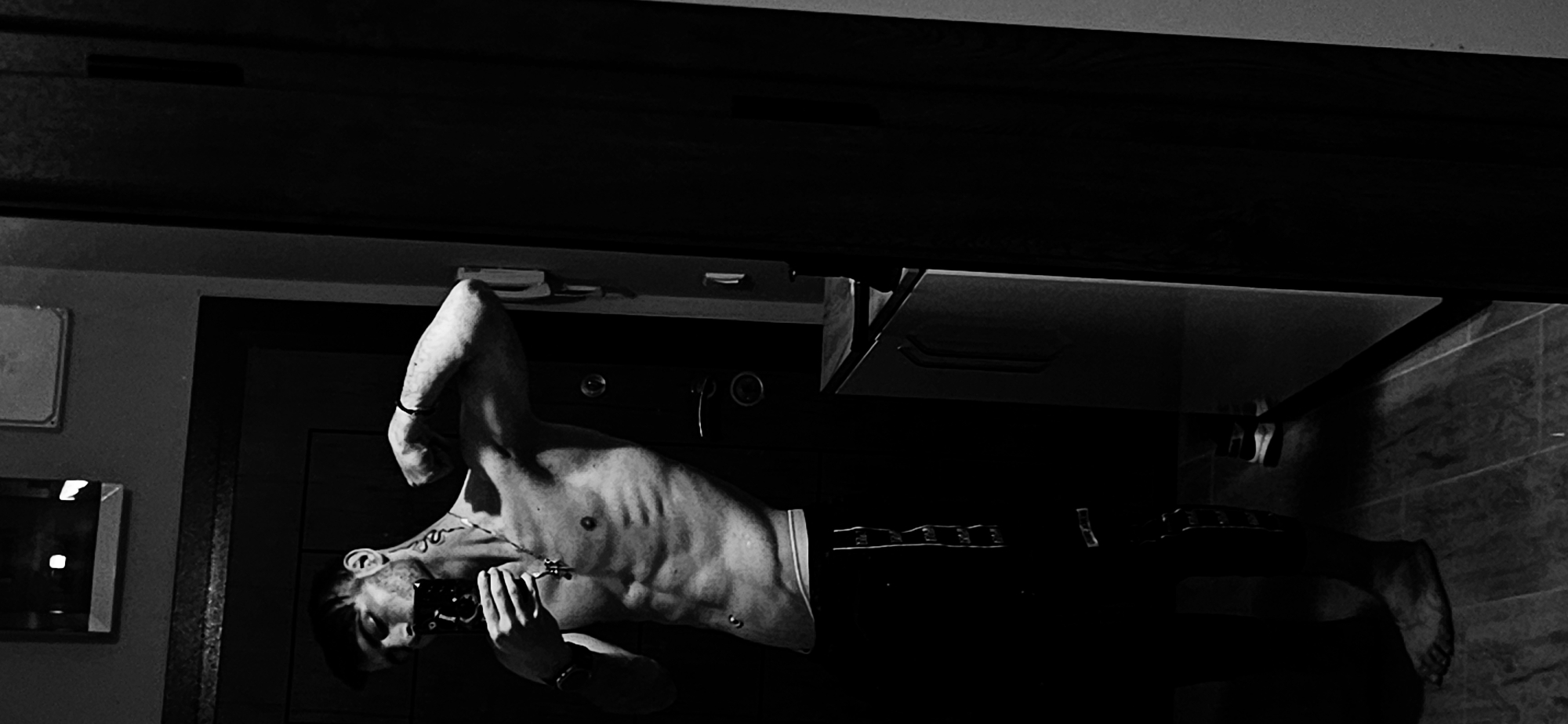 A black and white, flipped image features a muscular shirtless man holding a rectangular object in his right hand. He has a bracelet on his left wrist and a necklace. The background appears to be a bathroom setting. To the left, a mirror is visible on the wall, along with door and cabinet. The room seems to be dimly lit. The image location cannot be determined as Plovdiv, Bulgaria.