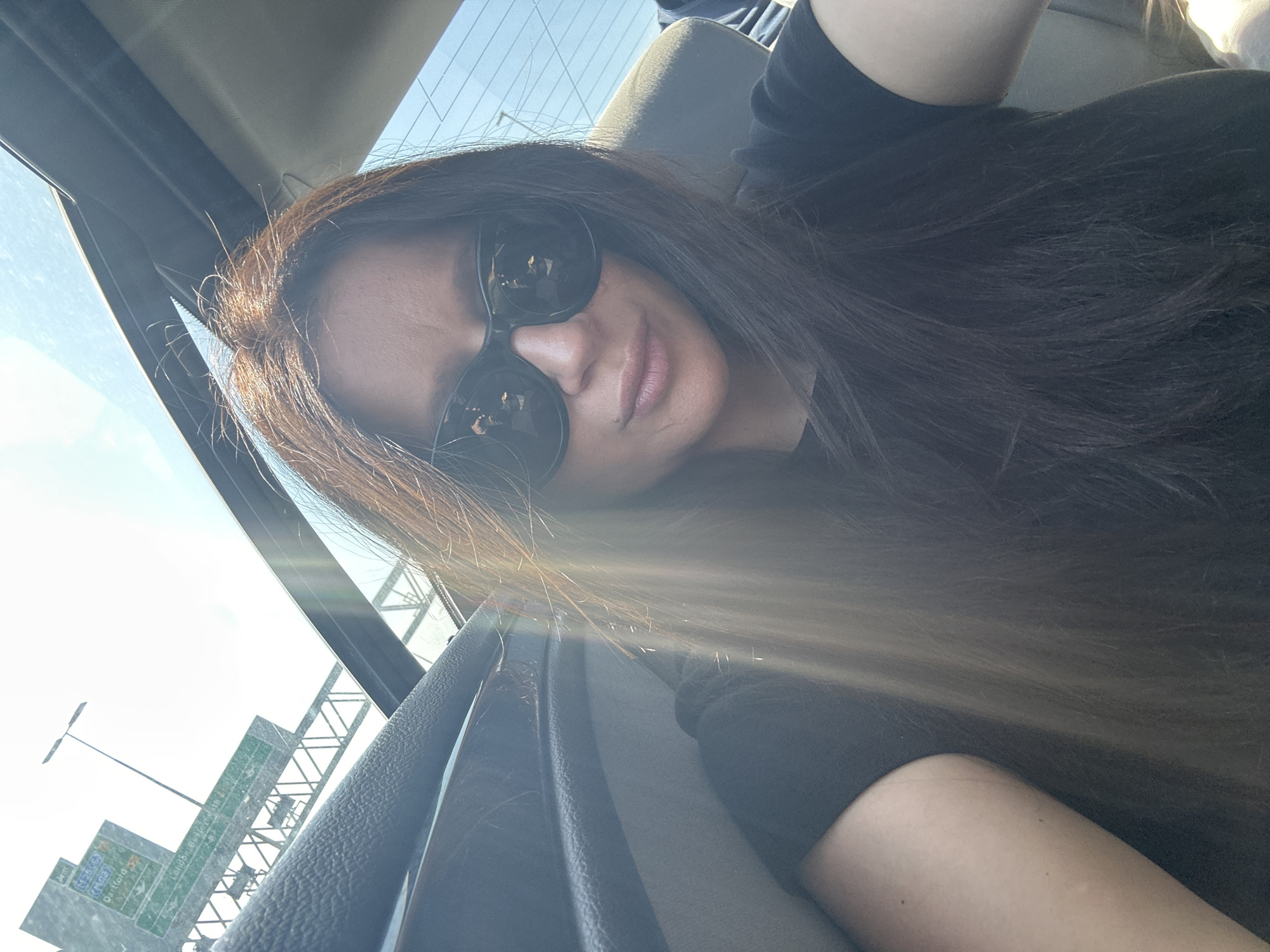 A woman with long brown hair and sunglasses is taking a selfie in a car. The background shows a highway sign indicating directions towards Oxford and London Bayswater Road. The location is Unknown, Unknown.