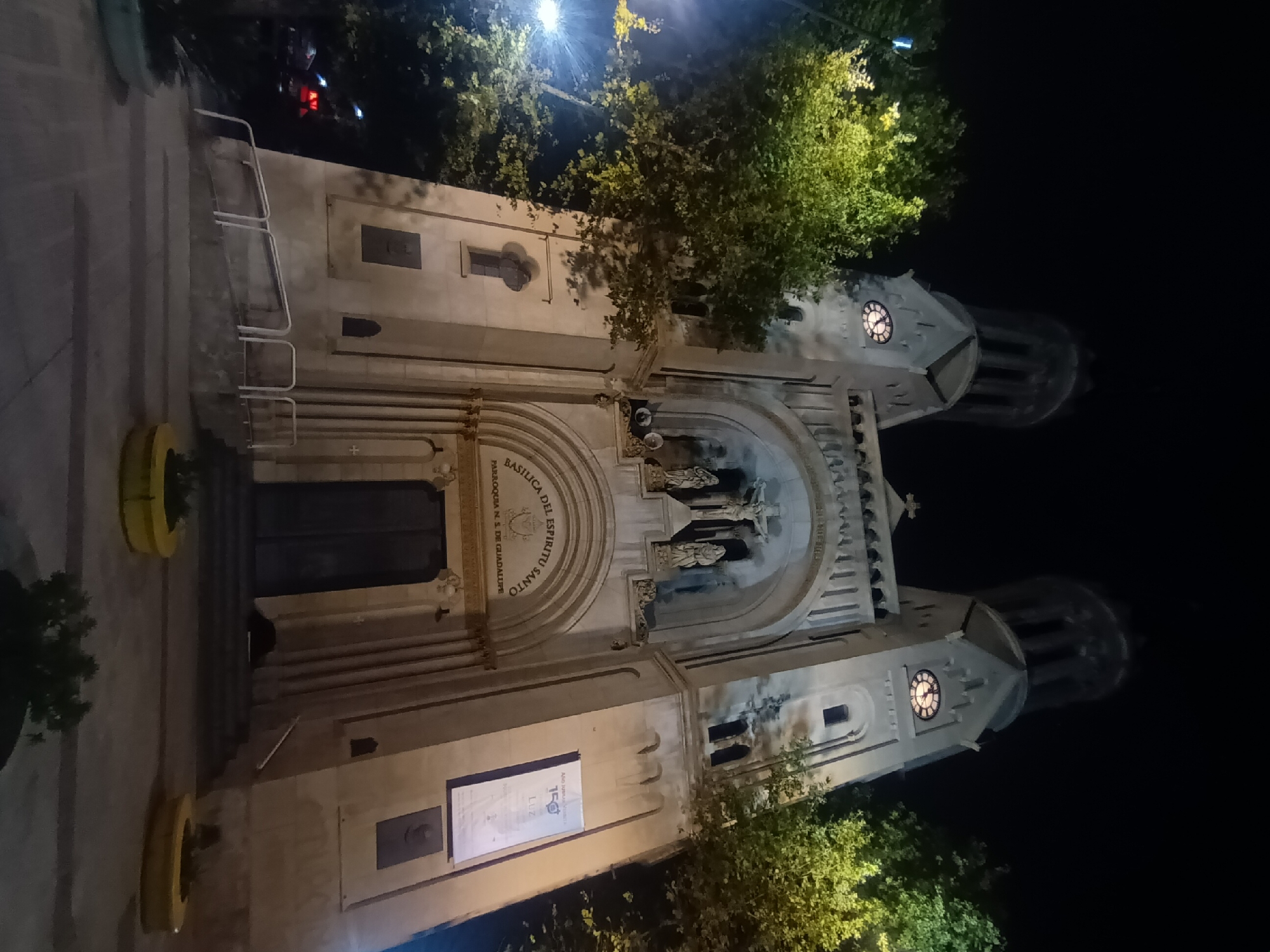 A nighttime exterior shot of the "Basilica del Espiritu Santo" in Buenos Aires, Argentina. The architecture is visible with its twin towers and decorative facade. The scene includes street lighting, and the basilica appears illuminated against a dark sky. Trees are visible, partially obscuring elements of the building. The perspective is from a street-level vantage point.