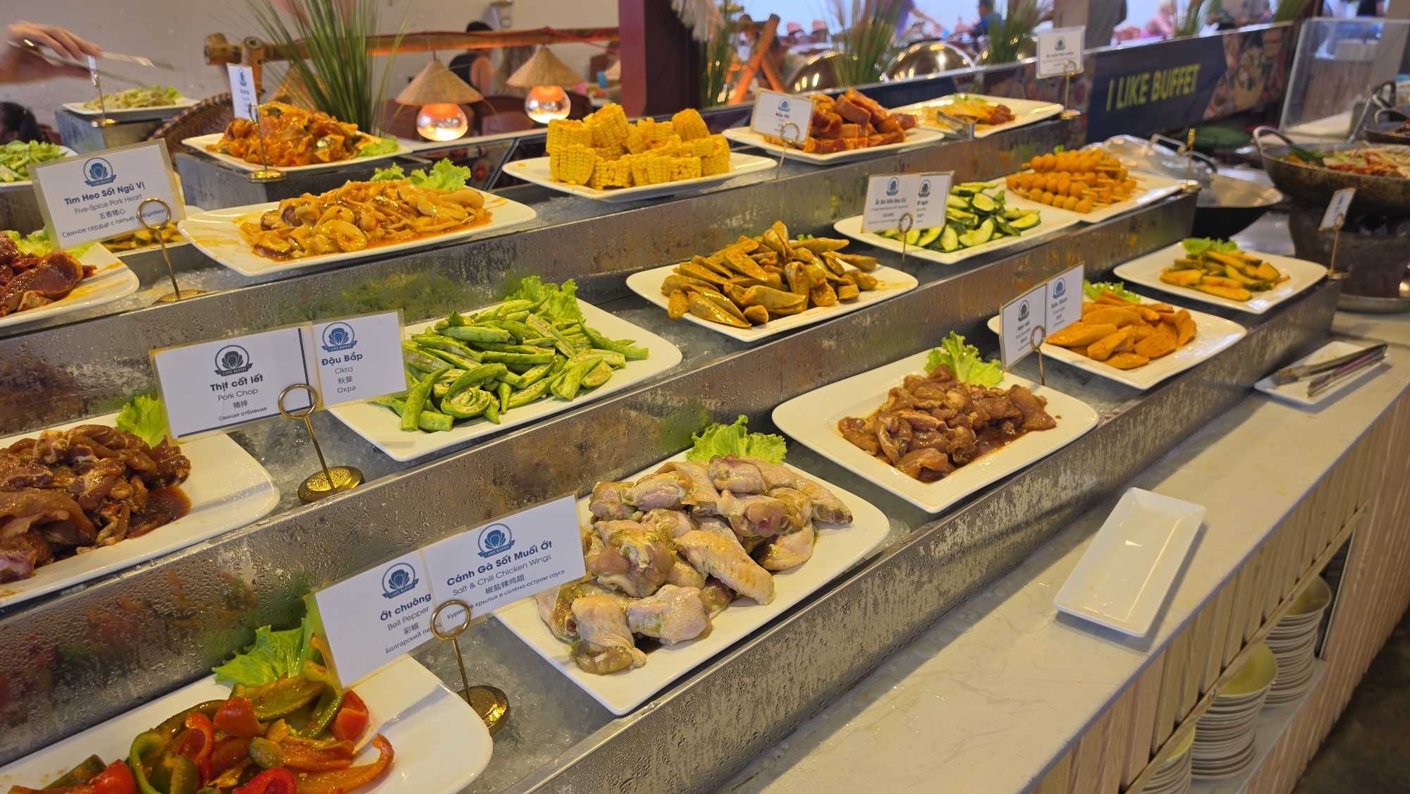 The image shows a buffet restaurant in Nha Trang, Vietnam, showcasing a variety of Asian dishes. The buffet is set up on a tiered stainless steel counter, with many dishes served in white rectangular platters. The dishes include an assortment of meats, vegetables, and seafood. Some of the identifiable dishes are pork heart, pork chop, okra, sliced cucumbers, chicken wings in salt and chili sauce, and sauteed vegetables. The food is presented with decorative lettuce garnishes and small informational signs with Vietnamese text and English translations. In the background, there are thatched lamps and people browsing the buffet. The overall setting appears to be an indoor dining area with ambient lighting.