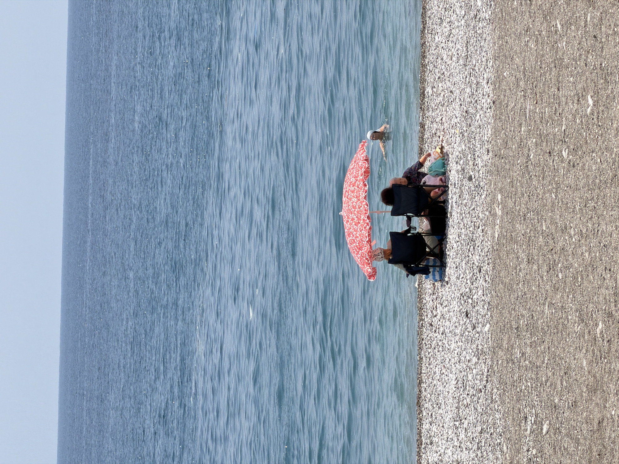 In Kemer, Turkey, on a sunlit day, two individuals are seated in camping chairs on a pebble beach under a red and white patterned umbrella. To the right of the umbrella, a third person is swimming in the clear blue water. The sea stretches to the horizon under a pale sky. A cooler bag and other items are visible near the seated individuals.