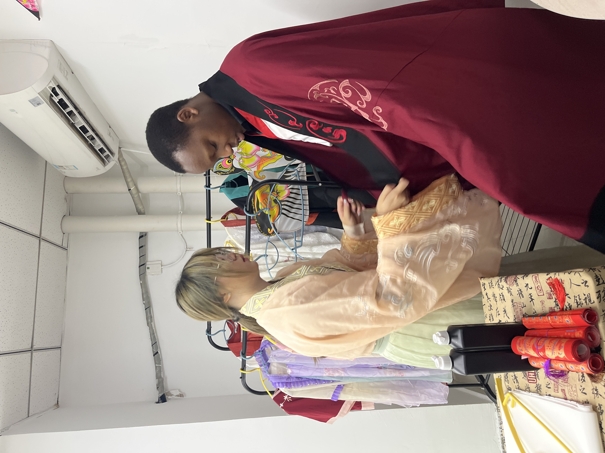 In an indoor setting, a person with dark skin and short hair is wearing a dark red robe with black and white trim. They are facing a person with fair skin and blonde hair, who is wearing a light orange robe with detailed embroidery on the sleeves. The fair-skinned person is standing by a clothing rack filled with garments in various colors. Adjacent to the clothing rack, there are rolled-up red scrolls and black ink bottles placed on a patterned fabric. The environment appears to be a dressing or preparation room, with a white ceiling, pipes, and an air conditioning unit visible. The scene does not include visual elements that permit identifying the specific location in Jiangjin, China.