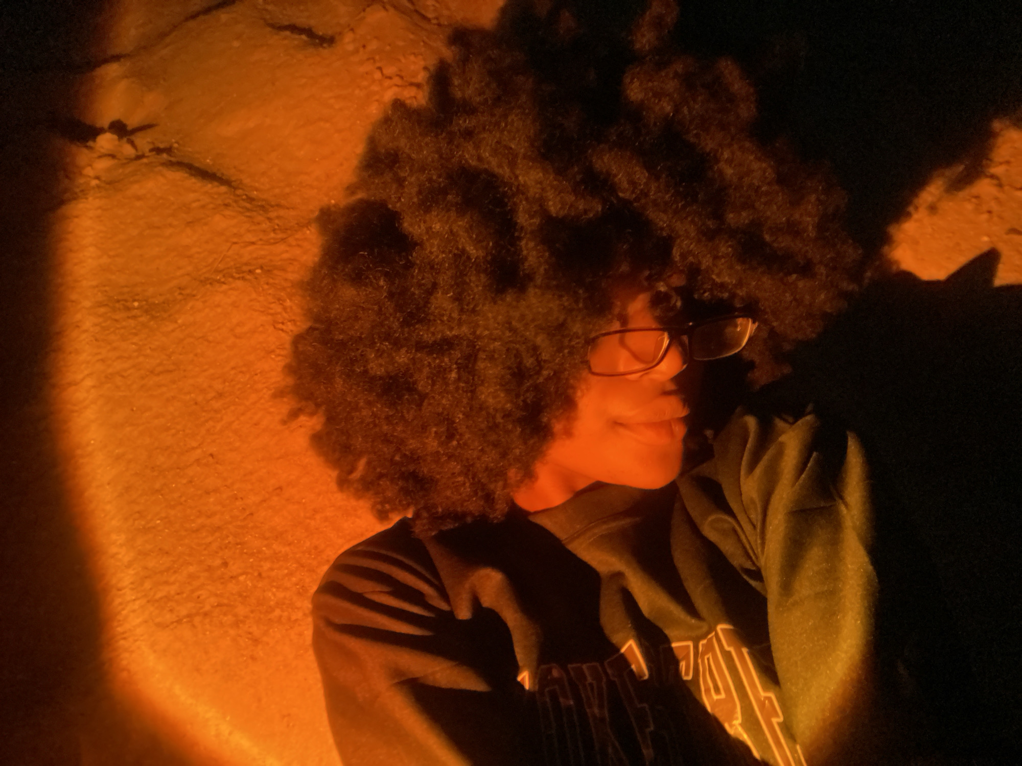 The image depicts one individual, a young person with dark, voluminous curly hair and dark-framed glasses, situated against a textured, orange-illuminated wall. The person's head is tilted slightly down and to the viewer's left, with eyes appearing closed or downcast, and a faint smile on their lips. They are wearing a dark long-sleeved garment, possibly a sweatshirt, with a partial, faint light-colored design visible on the chest. A strong orange light source illuminates the left side of the scene, casting a warm glow on the individual's face and hair, and the immediate background. The right side of the individual and the background are in deep shadow. The background surface, visible primarily on the left, exhibits a rough texture and subtle linear imperfections or cracks. The scene is located in Iranla, Nigeria.