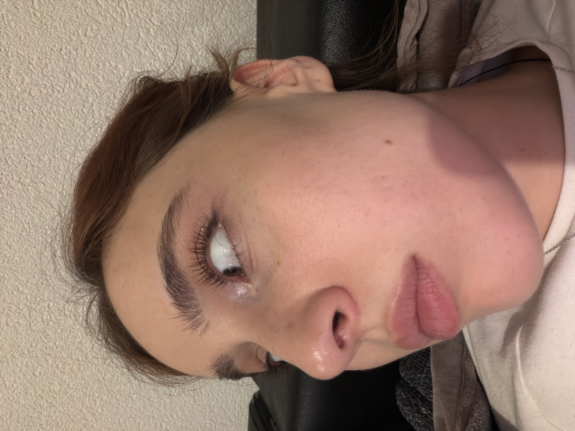 The image shows a close-up of a woman's face, angled slightly to the side. Her hair is brown, and her skin tone is fair. She has prominent eyebrows and long eyelashes. Her lips are slightly parted. The background is a textured off-white wall and what appears to be a dark-colored chair or seat. The environment does not provide enough evidence to place her in Strasbourg, France.
