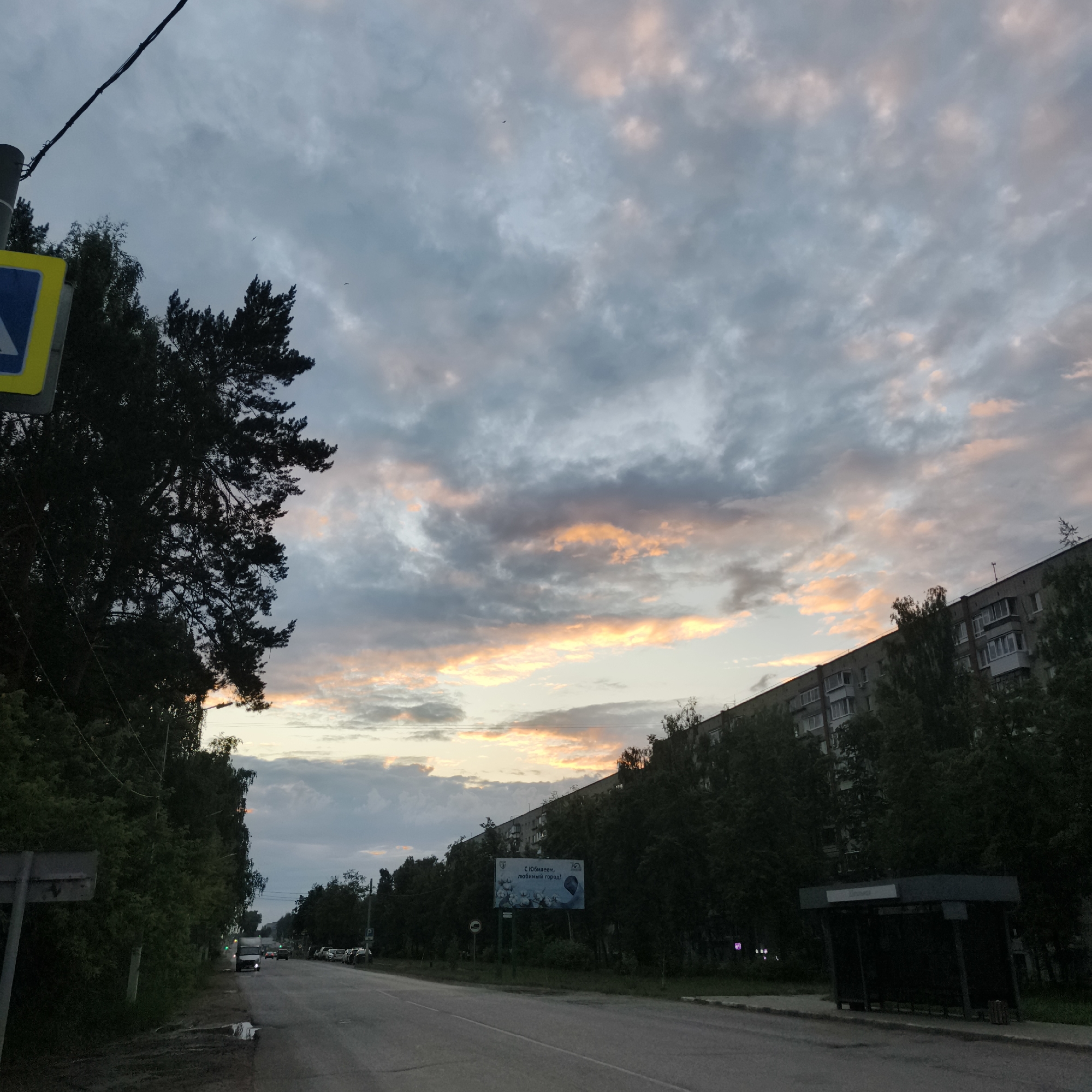 This media file depicts a street scene in Chaikovsky, Russia, likely during the evening given the dusky sky. The sky is filled with dramatic clouds, tinged with soft oranges and pinks from the setting sun, contrasting with the cooler blues and grays of the overcast sky.
The foreground shows a paved road with cars visible, suggesting ongoing traffic. On the left side of the road, there is dense greenery, including trees and bushes, and a pedestrian crossing sign. A tall, dark evergreen tree dominates the left side of the frame. On the right side of the road, a multi-story apartment building stretches into the distance, partially obscured by trees. A bus stop shelter is also visible on the right sidewalk.
A billboard is prominently displayed on the right side, advertising "С Юбилеем, любимый город!" which translates to "Happy Anniversary, beloved city!". This text indicates a celebratory event or anniversary for the city.
The overall mood is calm and slightly melancholic, characteristic of a twilight scene. The lighting suggests it's either dusk or dawn. The presence of buildings, road infrastructure, and vehicles places the scene in an urban or suburban environment.