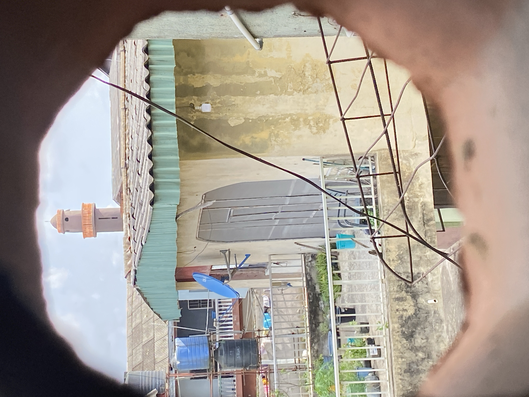 The image presents an urban scene in Lagos, Nigeria, viewed through an obscured circular opening. A mosque minaret is visible in the distance against a cloudy sky. The immediate foreground consists of densely packed residential buildings with corrugated metal roofs. Water tanks, satellite dishes, and electrical wiring are prominently displayed on rooftops and balconies. The buildings have peeling paint, suggesting weathering and age. The overall environment appears cluttered and densely populated. No people are directly visible.