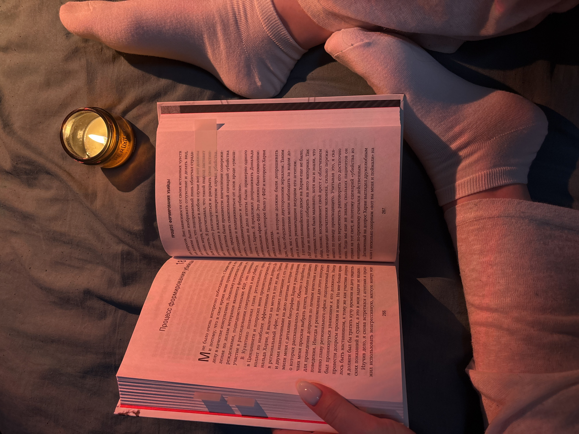 A person wearing white socks is lying in bed reading a book by candlelight. The book is open to pages 266 and 267. The text is in Russian and appears to be about the process of forming a murderer. The title of the book page 18 is "Процесс формирования, убийцы" The candle is in a gold-colored glass holder. The bedsheets are dark.