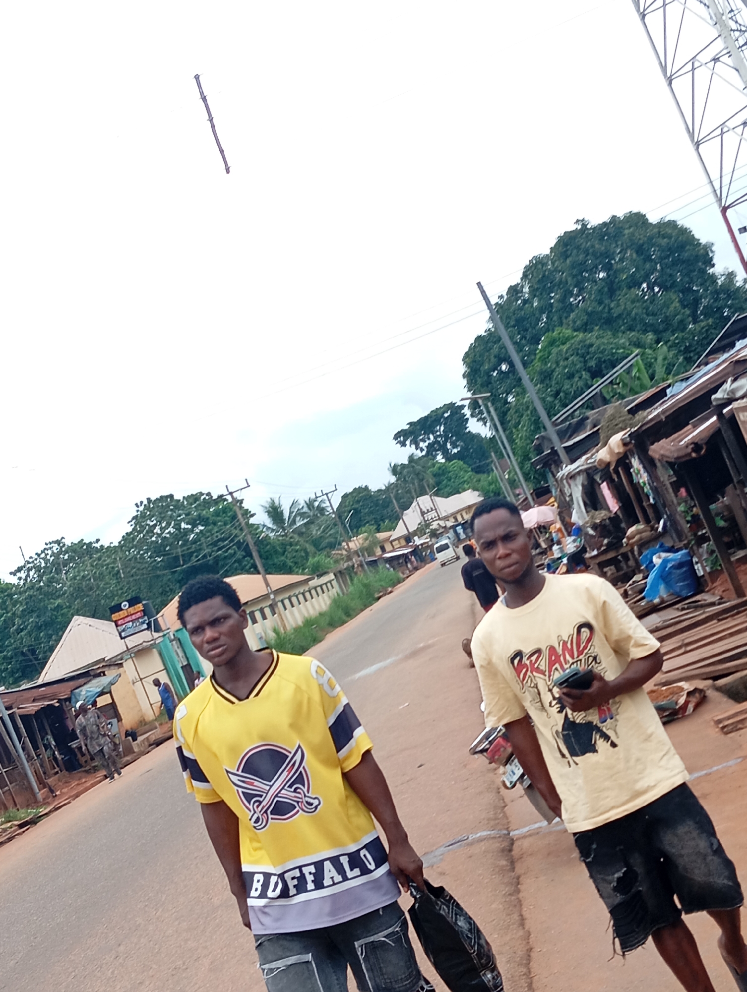 In the city of Nsugbe, Nigeria, two young men are captured in a candid moment on a dusty, paved road. The man on the left is wearing a vibrant yellow jersey with a "Buffalo" emblem and intricate designs. He carries a dark patterned bag. The man on the right is sporting a light yellow t-shirt with a graphic print that includes the word "BRAND" and dark, ripped denim shorts. He is holding a smartphone in his right hand. The backdrop reveals a street lined with local shops constructed from corrugated iron and wood, displaying an assortment of goods. A few motorbikes are visible, hinting at the mode of transportation in the area. Trees and vegetation frame the scene, with a radio mast and power lines overhead suggesting the presence of infrastructure. The sky is overcast, indicating a cloudy day. The general atmosphere appears to be that of a bustling local market or street.