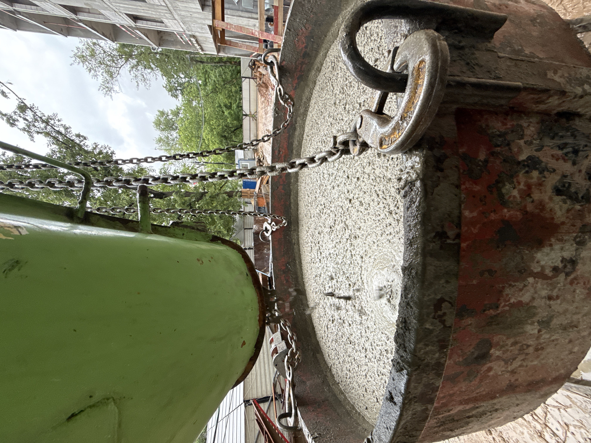 An overhead shot captures a construction site in Moscow, Russia, where liquid concrete is being poured from a large green hopper into a heavy-duty red metal basin. The concrete, a pale gray slurry, fills the basin to the brim, with droplets actively falling from the hopper's opening. Several thick metal chains suspend the hopper, with one chain attached to a large, U-shaped metal clasp secured to the rim of the basin. The basin itself appears worn and stained with dried concrete and mud. In the background, trees and an unfinished multi-story building under a cloudy sky indicate an active construction project. The ground is uneven and appears to be made of dirt and mud, consistent with a construction environment.