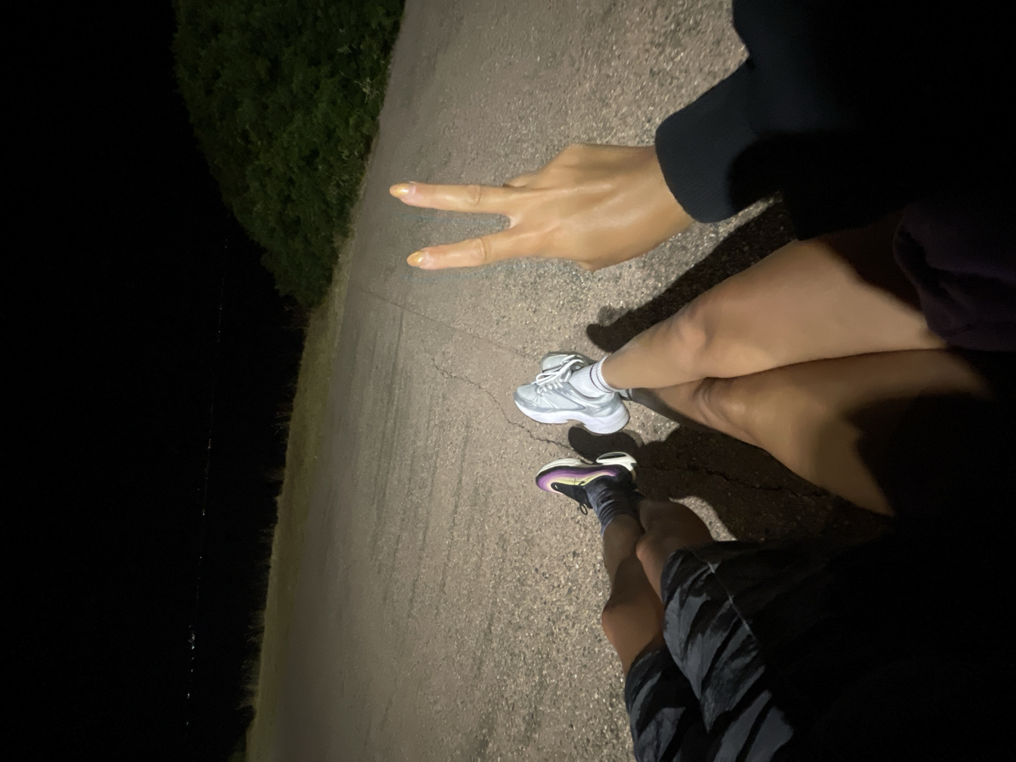 A low-angle, close-up shot shows two people's legs and one hand as they sit on a paved surface at night. The person on the left wears black and grey patterned shorts and dark socks with multi-colored athletic shoes. Their legs are bent at the knee, with the feet resting on the ground. To the right, a person wearing white athletic socks and grey athletic shoes also has their legs bent at the knee, with their feet on the ground. Between the two sets of legs, a right hand is visible, with the index and middle fingers raised in a peace sign. The hand has painted nails with a peachy-gold color. The skin tone of the people's legs suggests they may be of different ethnicities. The paved surface appears to be a path or road, with faint lines and pebbles visible. In the background, a dark, blurry expanse suggests a body of water with distant lights, possibly indicating a city or town. To the right, a dark, dense green bush or hillside is visible. The overall lighting is dim, with light sources creating shadows on the paved ground. The location is the Municipal Unit of Kassandra, Greece.