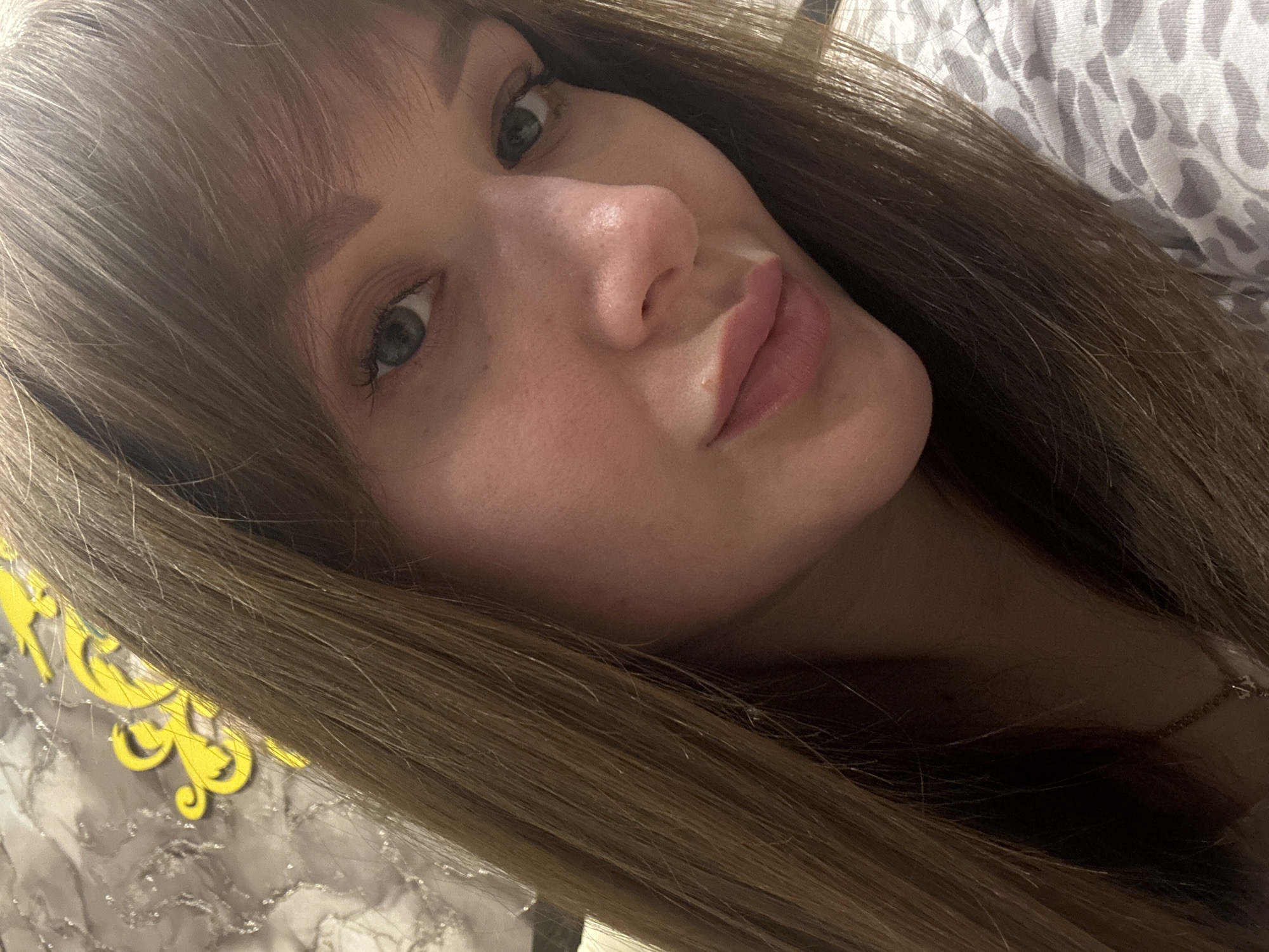 A close-up, slightly tilted portrait of a young woman with light brown, straight hair and bangs is displayed. She has fair skin, green eyes, and full lips. She is wearing what appears to be a light-colored patterned top. Part of a yellow decorative object is visible in the lower left corner, possibly on a surface with a marble-like pattern. A delicate necklace is also visible around her neck.
There is no verifiable evidence in the image to confirm a location in Chaikovsky, Russia.