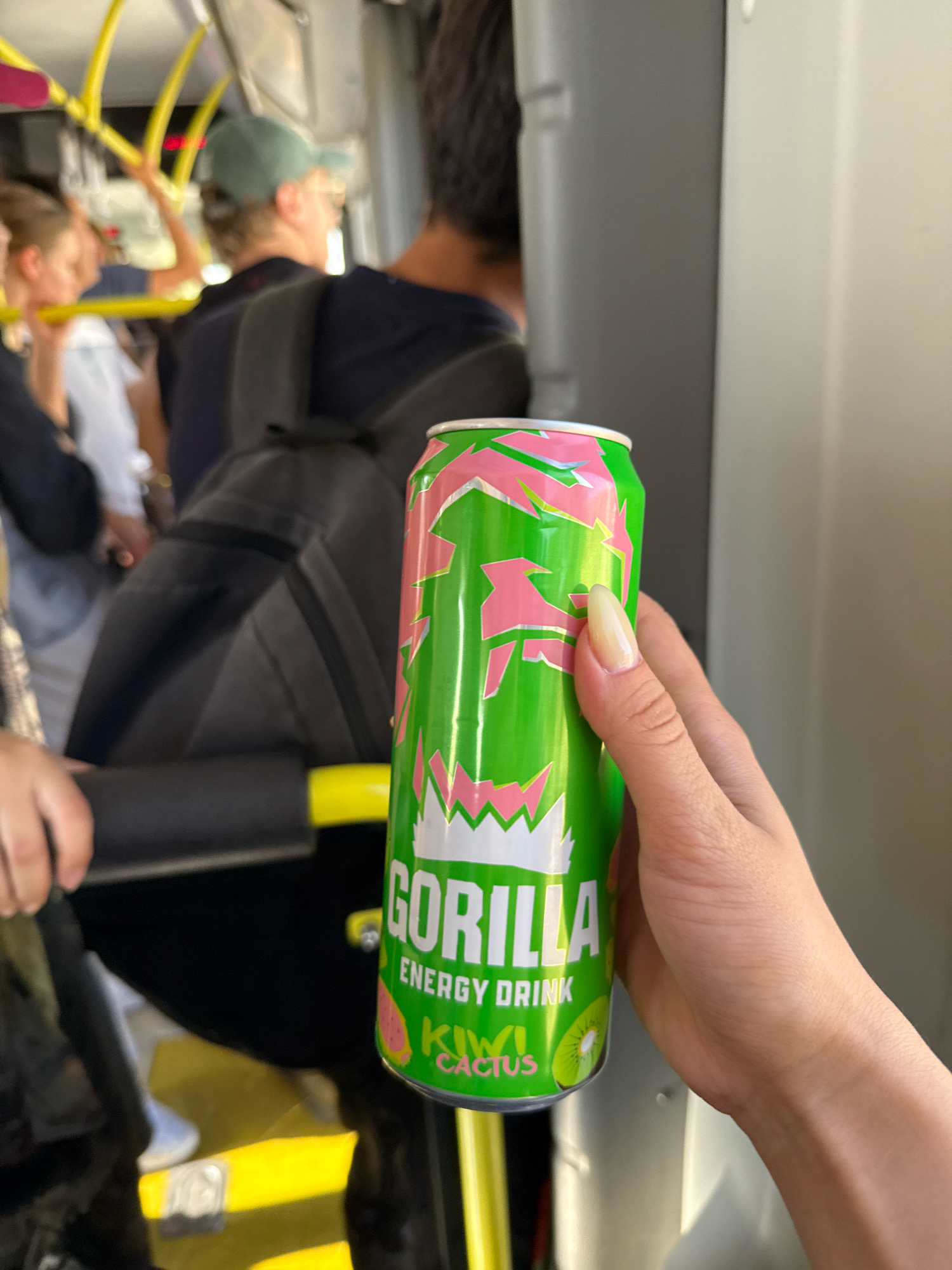 The image depicts the interior of a bus in Zarechnoe, Kyrgyzstan, focusing on a hand holding a green can of "Gorilla" energy drink. The can features pink and white designs, with the text "GORILLA ENERGY DRINK" and "KIWI CACTUS" clearly visible. In the background, passengers are visible, including a person wearing a green baseball cap and another with a black backpack. Yellow poles and grab handles, characteristic of bus interiors, are also present. The lighting suggests it is daytime.