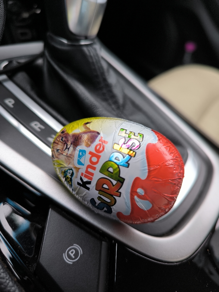 A Kinder Surprise egg, still sealed in its multi-colored foil wrapper, rests on the dark gray center console of a vehicle. The wrapper displays a red lower half, a white middle section with "Kinder Surprise" text, and a yellow upper section featuring a partial image of a feline animal. The foil shows minor creasing, notably on the red portion. In the background, an automatic transmission gear shifter with a black leather boot and silver top is visible. Adjacent to the shifter, a gear selection display indicates "P" and "R". To the lower left of the Kinder egg, an electronic parking brake button marked with a circular "P" symbol is present on the console. A blurred light-colored car seat is partially discernible in the right background. No individuals, explicit actions, or direct interactions are depicted. The specific location within Illintsi, Ukraine, cannot be determined from the visual information provided.