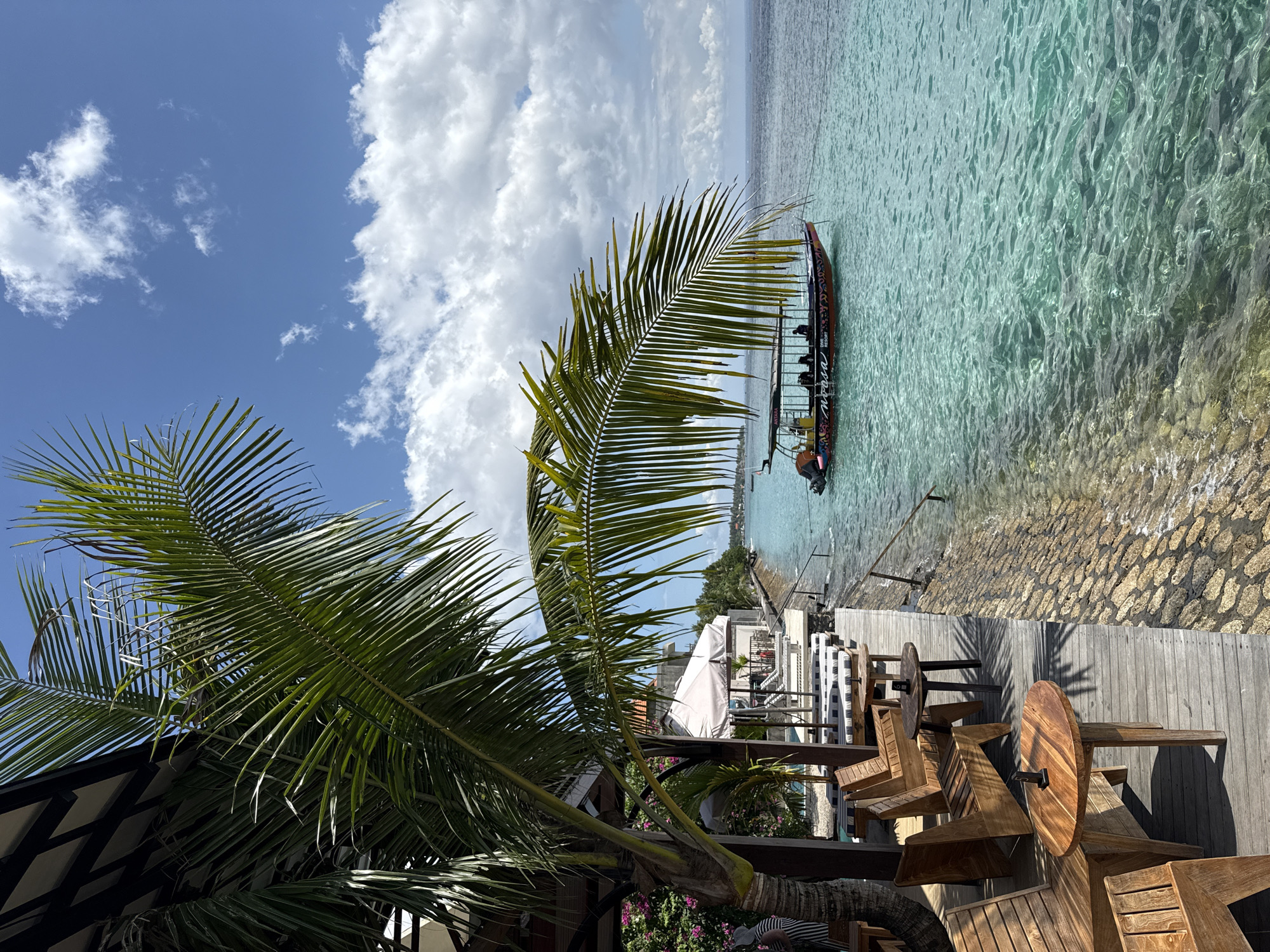 The image shows a tropical scene in Nusa Penida, Indonesia, featuring a wooden deck area with tables and chairs overlooking a clear, turquoise body of water. A boat is docked near the shoreline. Palm trees are in the foreground. The sky is blue with scattered white clouds. The decking extends to the rocky shoreline.