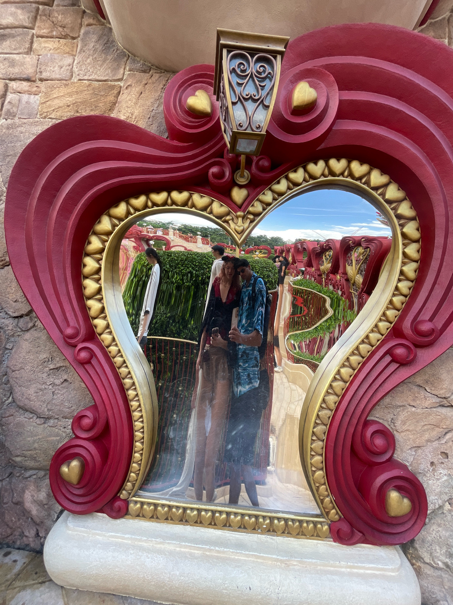 A heart-shaped mirror, adorned with red and gold detailing, reflects a couple taking a selfie. The mirror is framed by an ornate red structure with gold hearts. Behind the couple, a park-like setting with red structures, green hedges, and a bright blue sky is visible. The mirror is placed on a stone surface. The location is Shanghai Disneyland in Pudong, China.