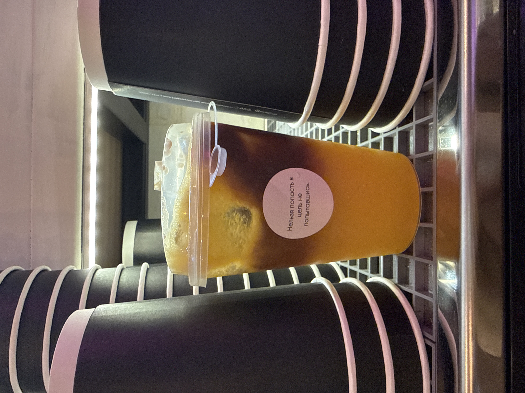 The image, captured in Khimki, Russia, displays a clear plastic cup holding a layered cold beverage, likely within a refrigerated display case or a vending machine. The drink is horizontally oriented and shows a distinct separation: a dark brown liquid forms the upper layer, while a bright orange or yellow liquid constitutes the lower layer, with ice cubes visible throughout.
A circular, light pink sticker is affixed to the side of the plastic cup, featuring the Russian phrase "Нельзя попасть в цель не попытавшись," which translates to "You can't hit the target without trying."
The central cup rests on a light gray metal grid or rack. Above and below it, multiple black paper cups with white rims are arranged horizontally in rows, suggesting they are either stored for use or are part of a beverage dispensing system. A bright vertical light strip, possibly an LED, illuminates the scene from the left, while a reflective metallic surface is visible on the far right, indicative of the display's interior. Faint, unreadable white text is partially visible on one of the black paper cups near the top. The overall setting implies a self-service or commercial beverage environment.