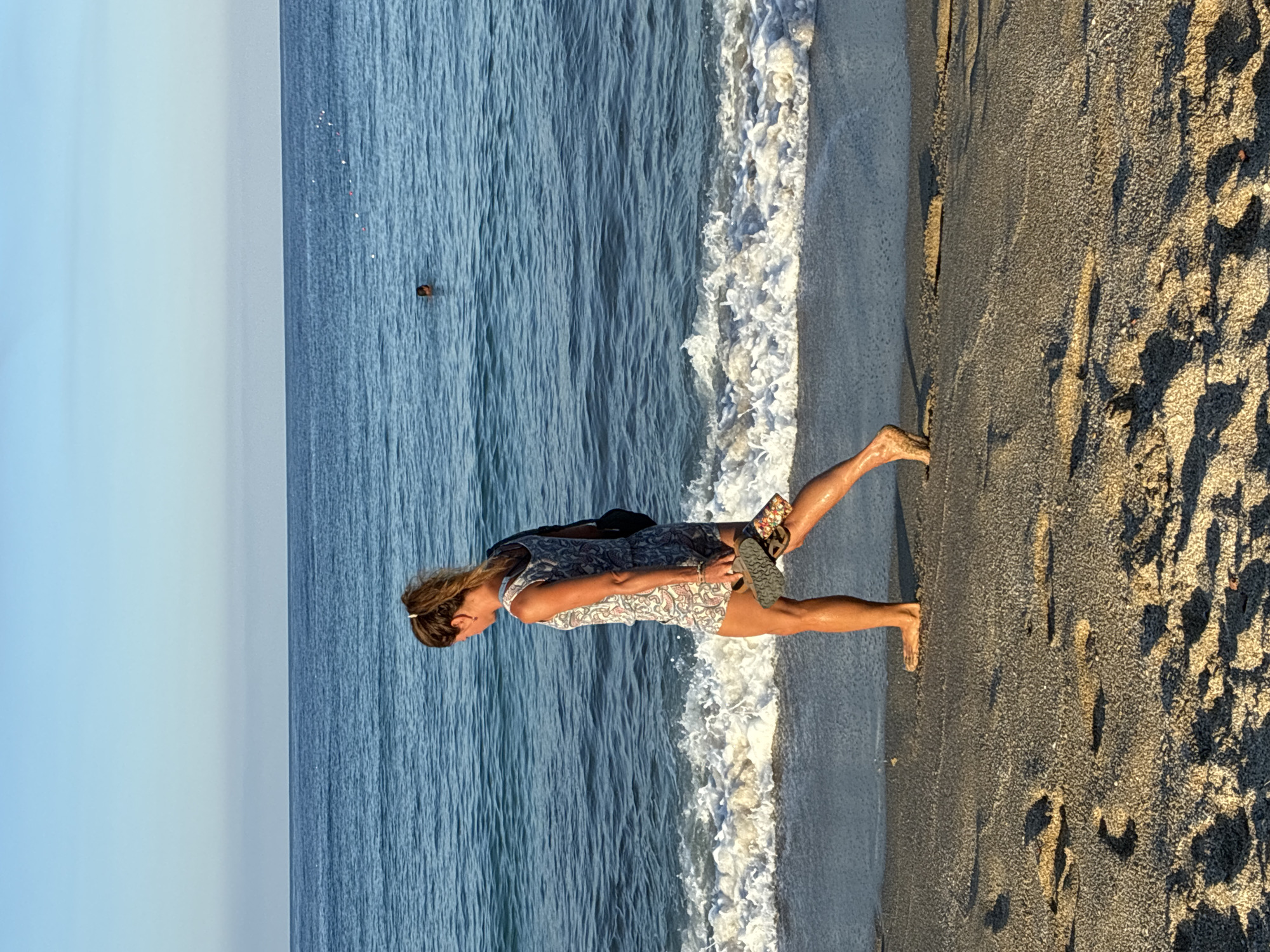 A woman is walking along the sandy shore of a beach. She is wearing a patterned short romper and carrying a pair of flip-flops in her right hand. Her left hand is by her side. A black bag is slung over her left shoulder. The ocean is calm with gentle waves lapping at the shore. In the distance, a person is swimming in the water. The sky is clear and blue. The scene appears to be in Alanya, Turkey, based on its coastal setting and general aesthetic.