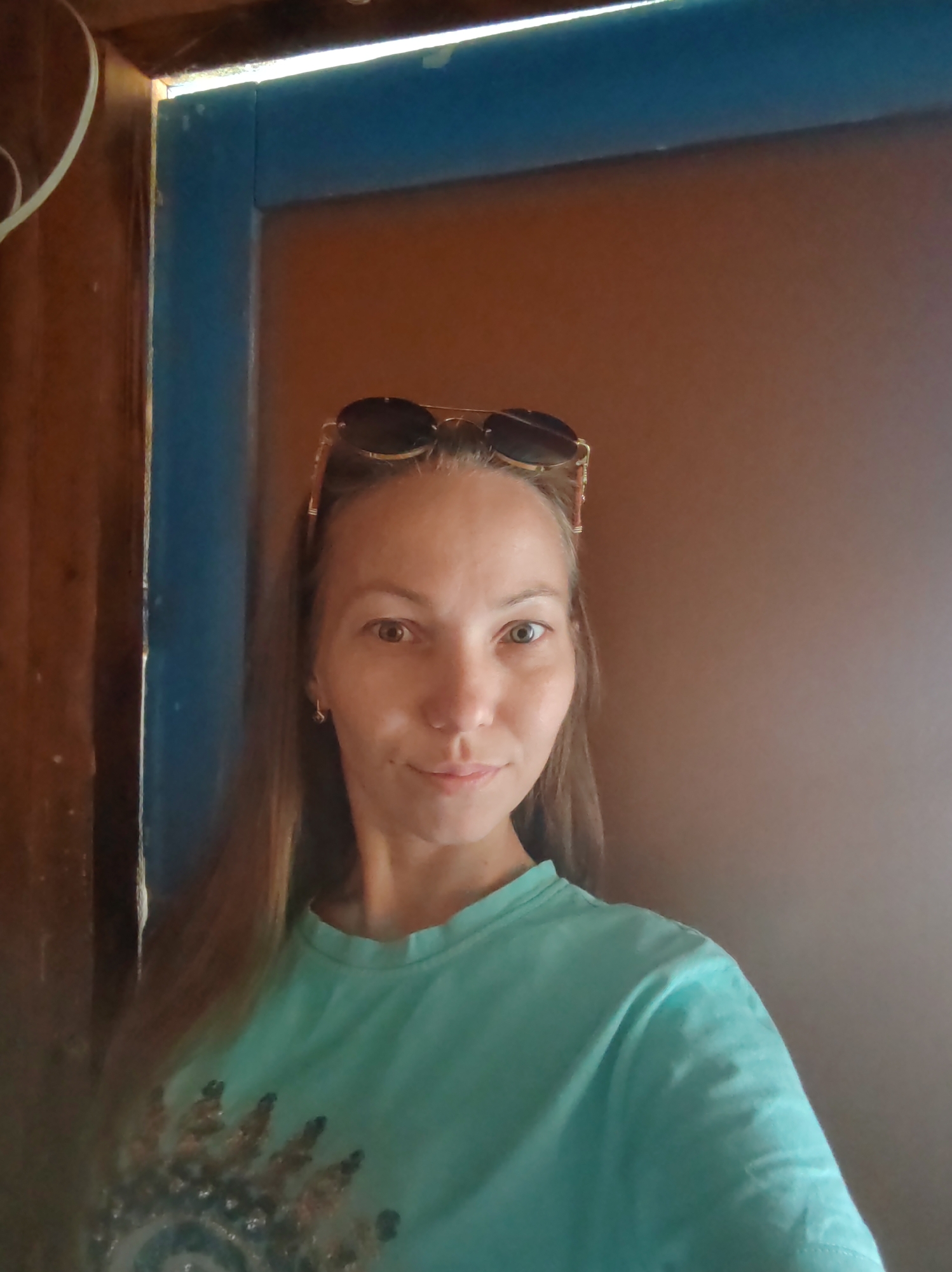 In the city of Chuchlem, Russia, a woman with long, brown hair and sunglasses perched on her head is captured in a medium shot. She is wearing a light blue t-shirt with a decorative emblem on the front. She is standing in front of a brown wall, with a wooden doorframe featuring blue accents to her left. The lighting suggests an indoor setting with natural light entering from the top left.