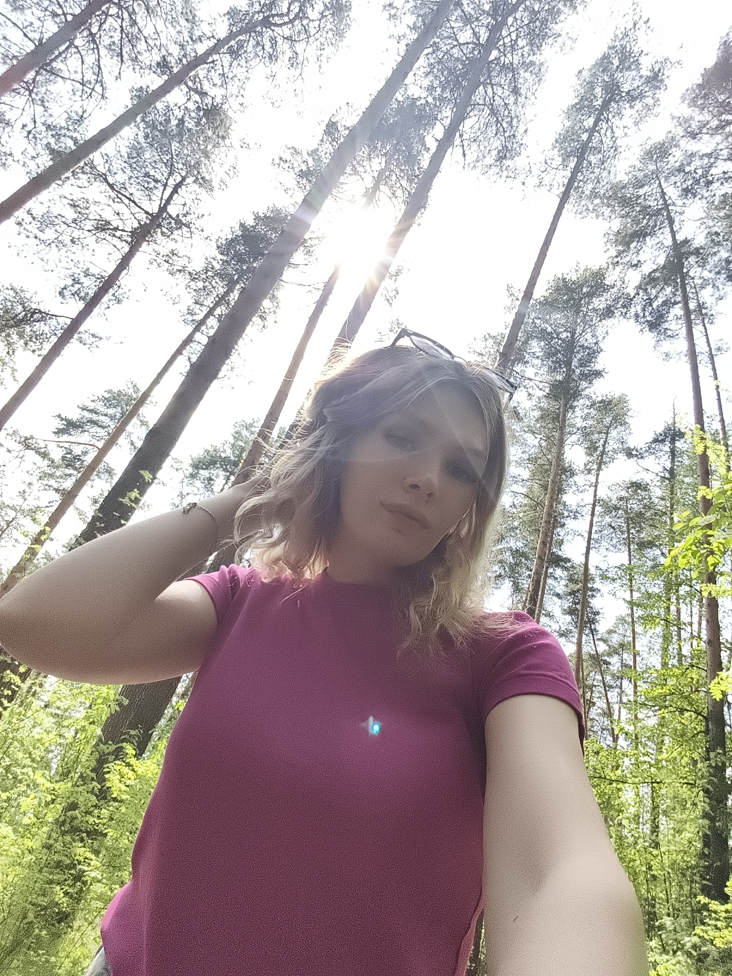 A low-angle, full-body selfie of a fair-skinned young woman with blonde, shoulder-length hair. She wears a pink t-shirt and has glasses resting on her head. Her left hand is raised to her hair. The background is a forest with tall, straight trees reaching upwards, their tops silhouetted against a bright sky, possibly the sun causing lens flare. Green foliage is visible at the lower edges of the frame. The location appears to be a wooded area, possibly on the outskirts of a city but there's insufficient information to place this photo in Izhevsk, Russia.