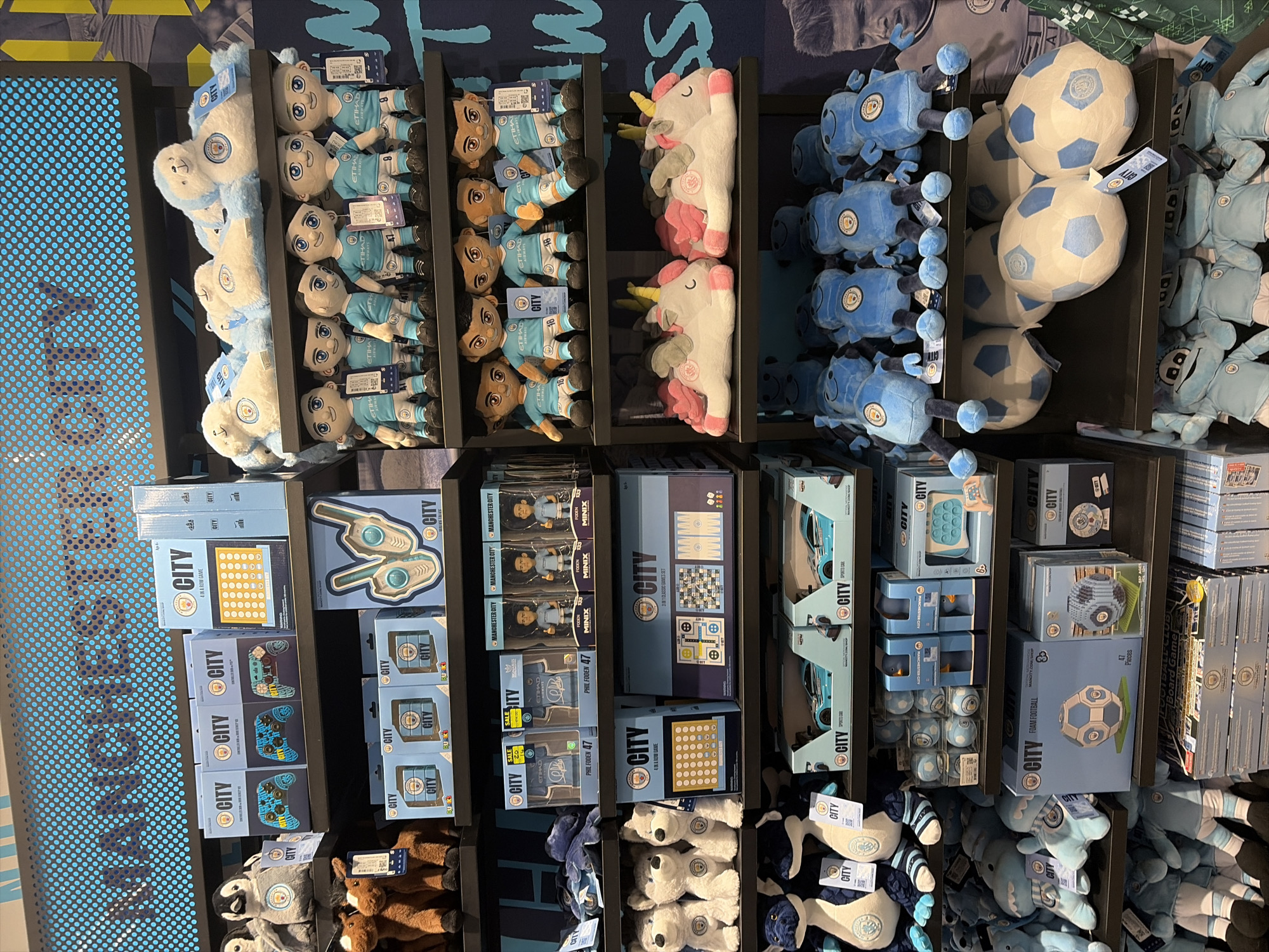 The image shows a shelf display of Manchester City football club merchandise. The items include plush toys of player figures in Manchester City kits, unicorn plushies, and football plushies. There are also boxed games such as a Connect Four game, a 3-in-1 classic game set, and a foam football set. The merchandise is arranged on black shelves with some items displayed in multiple quantities. A section of a blue perforated panel is visible to the left, which includes the words "Manchester City". The image appears to be taken inside a store selling Manchester City merchandise, located in the city of Manchester, United Kingdom.