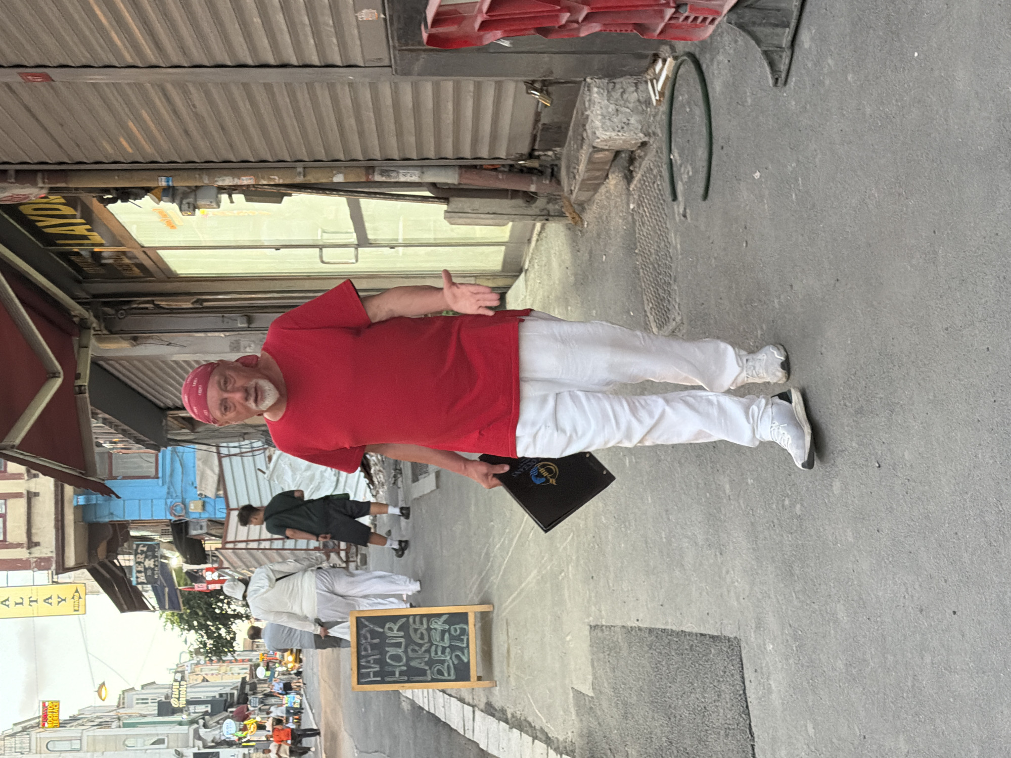 The image shows a man in a red t-shirt and white pants walking down a street in Istanbul, Turkey. He is wearing a red bandana and has a beard. He is holding a black folder in his left hand and has his right hand outstretched, palm up. He is walking on a sidewalk next to a building with metal shutters. A chalkboard sign that reads "HAPPY HOUR LARGE BEER 2£9" is visible on the left side of the image. Other people are walking in the background. The street appears to be a busy commercial area with shops and signs in Turkish.