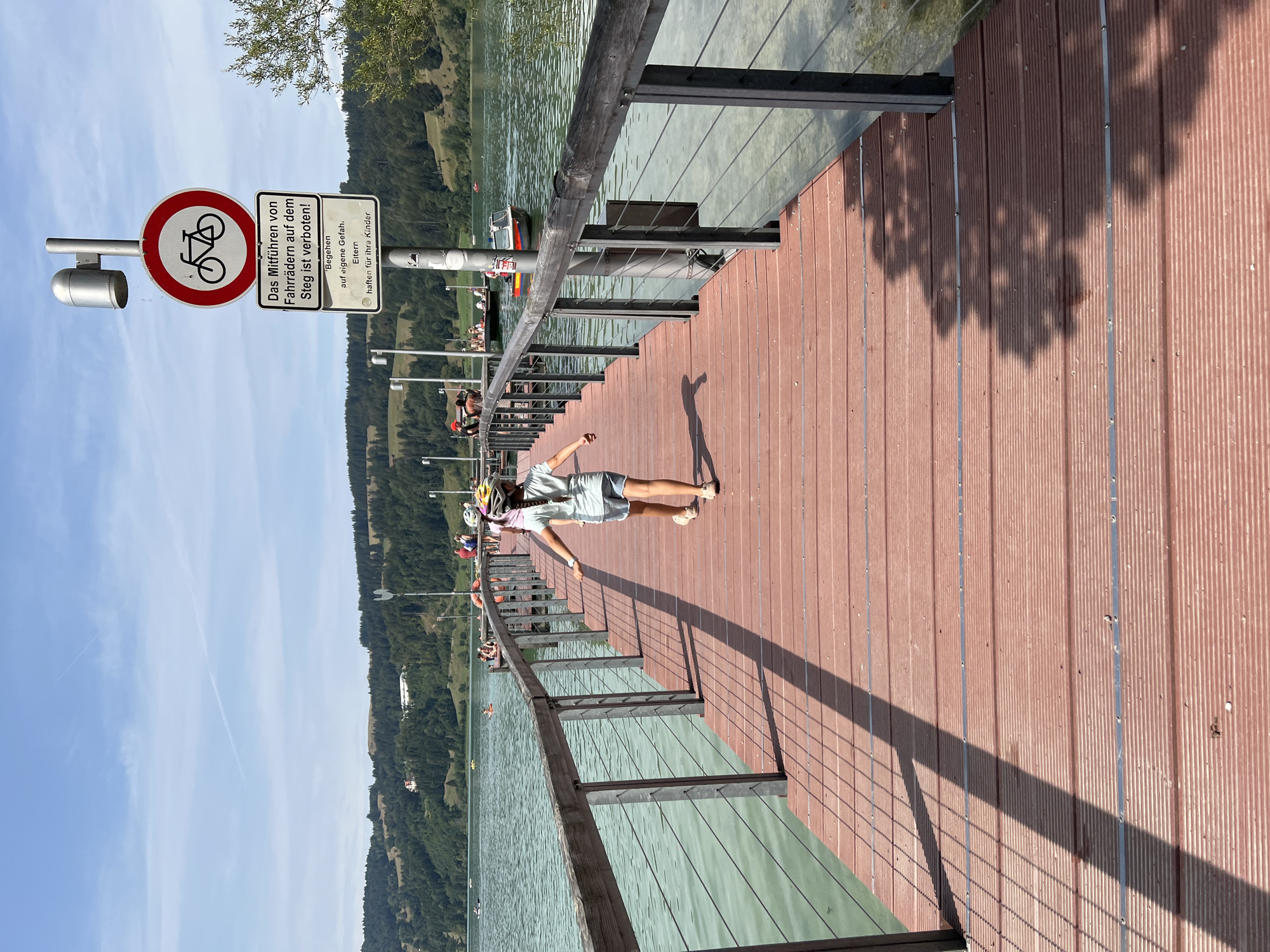 An eye-level, medium shot captures a wooden pier extending into a body of water, likely Alpsee Lake in Immenstadt im Allgäu, Germany, under a clear blue sky. A young girl, seen from behind, walks down the pier towards the camera, wearing a light blue t-shirt, denim shorts, and a yellow helmet. She has a pink braid and is wearing beige shoes. Her shadow stretches across the wooden planks of the pier. To the left of the pier is the calm, greenish-blue water of the lake, with a wooden railing along the pier's edge. In the distance, a tree-covered hillside forms the backdrop, dotted with buildings. A signpost on the pier displays a bicycle symbol crossed out, indicating that cycling is prohibited, and further text in German. Several other people are visible on the pier and in boats on the lake, suggesting a recreational setting.