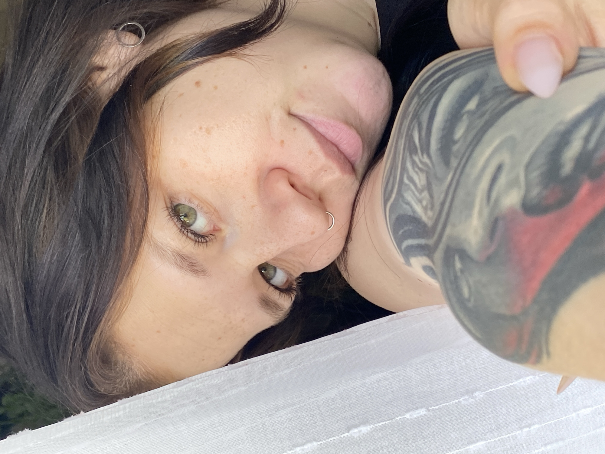A selfie photograph of a woman with fair skin, brown hair, and green eyes. She has freckles on her face and piercings in her ear and nose. Part of a tattoo is visible on her right arm. The background consists of what appears to be a white sheet or fabric with faint greenery visible in the blurred distance. It is impossible to confirm the location as Chaikovsky, Russia, from the visual information provided in the image.