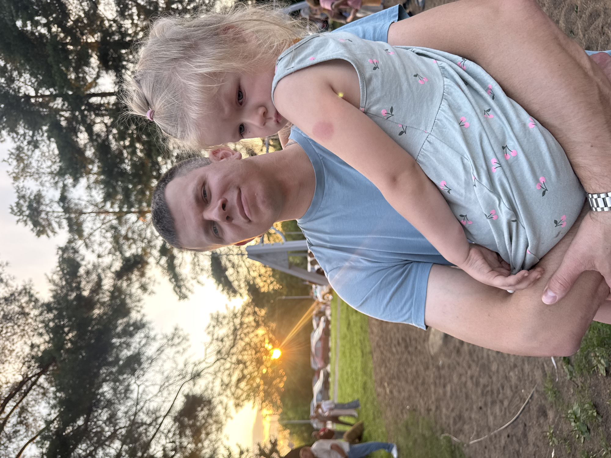 An outdoor, eye-level, medium shot features a man holding a young girl. The man, wearing a light blue t-shirt, faces forward with a neutral expression. He holds the girl across his chest; she faces left with a sad expression, her blond hair pulled back with a pink elastic. A red mark is visible on the girl's upper left arm. The background shows trees with the sun setting behind them, casting a warm glow. In the middle ground, cars are parked in a grassy area, with several people walking around. The foreground consists of a dirt and grass area, possibly a playground. The scene appears to be in a park or recreational area.