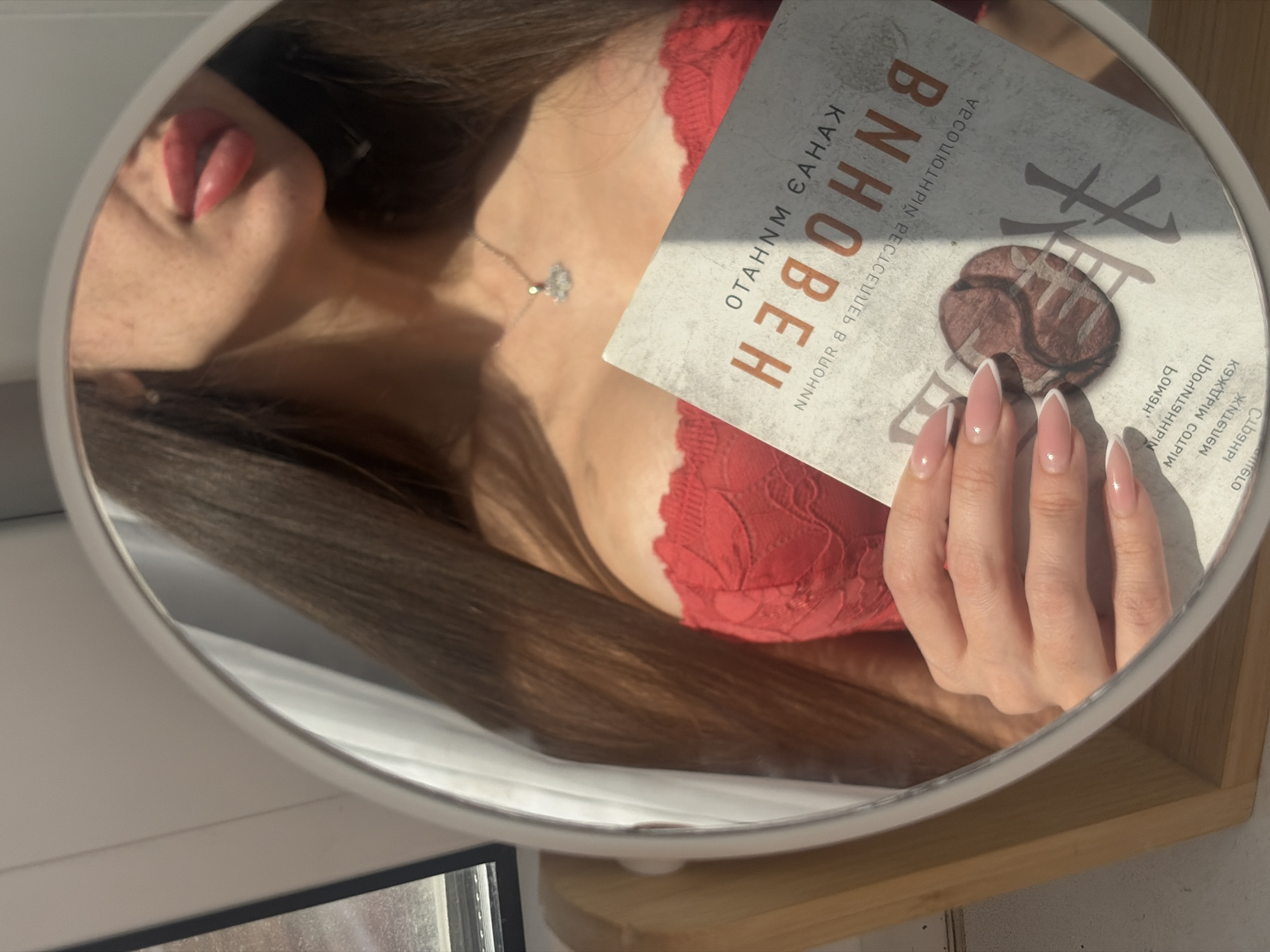 An unidentified female subject is depicted indoors, reflected in an oval-shaped mirror with a white frame, positioned on a light-colored wooden stand. The location is Saint Petersburg, Russia. The subject has long, dark brown hair and is wearing a red lace bralette or top. Her lips are made up with red lipstick, and a silver-colored necklace with a small pendant is visible around her neck. Her left hand, featuring manicured nails with pink bases and white French tips, holds a book. The book's reflected title is "ВНОВЕН" (Vnoven), identifying it as "ВНОВЕННЫЙ" (The Newcomer/Entered) by author Stanislav Vostryakov, with a circular, brown, stylized symbol on the cover. A window and white curtain are partially visible in the reflection to the left of the subject. Natural light from the window illuminates the scene, casting shadows across the book cover.