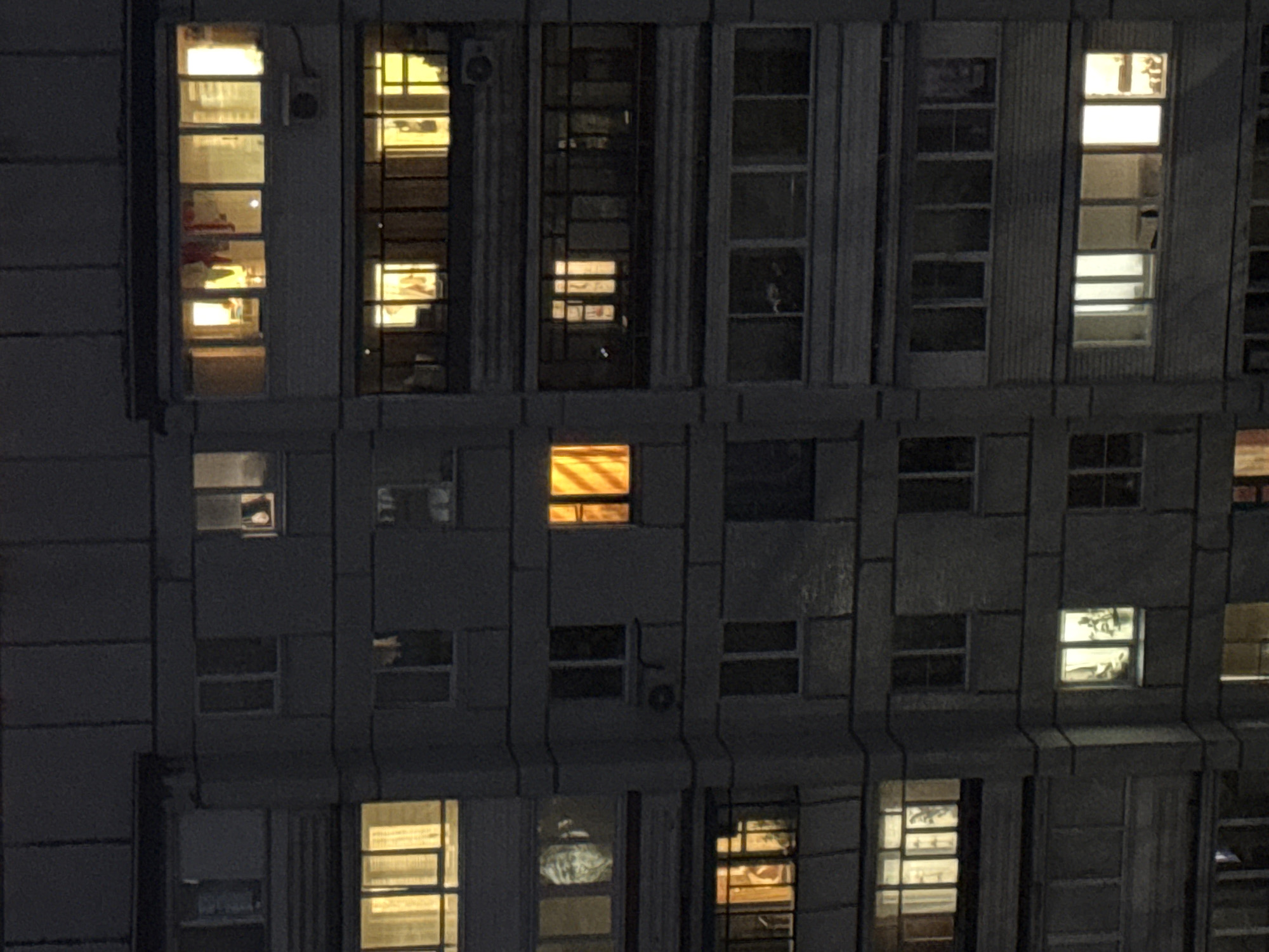 The image shows a section of a tall building at night, with multiple illuminated windows. Some windows reveal details of the interiors, suggesting apartment residences or office spaces. A few of the windows have furniture and other objects inside the room. The building exterior is dark and appears to be constructed of concrete or stone. The style of architecture might be categorized as Soviet-era apartment complex. There is no indication of this location being in Chaikovsky, Russia.