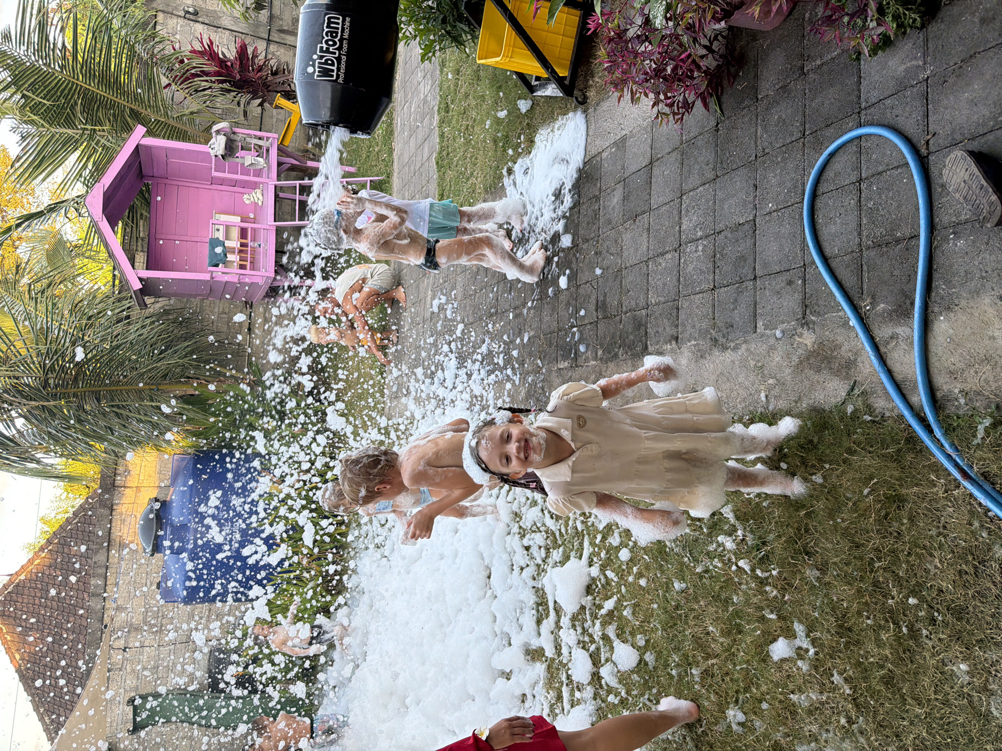 This image captures a vibrant outdoor foam party in Kuta Selatan, Indonesia. A black "WB Foam Professional Foam Machine" is actively dispensing copious amounts of white foam across a grassy area and an adjacent paved patio.
Multiple children are joyfully engaged in playing amidst the foam. Prominently in the foreground, a girl with braids, wearing a light-colored dress, smiles at the camera, her face and body covered in foam. Behind her, a boy in blue swim trunks and another child are also standing in the foam, close to the machine. Further back, another child is visible playing in the thick foam. Part of an adult's leg in red shorts is visible at the bottom of the frame.
The setting is bright and sunny, indicating daytime. Tropical elements include lush green grass, tall palm trees on the left, and red-leafed plants in the background. A pink wooden playhouse stands near the palm trees. The ground consists of grey square pavers on the right, where a coiled blue garden hose is also present. In the background left, a building with a tiled roof and a blue refuse bin can be seen. The children's expressions suggest happiness and fun during the event. The text "WB Foam Professional Foam Machine" is clearly visible on the black device.