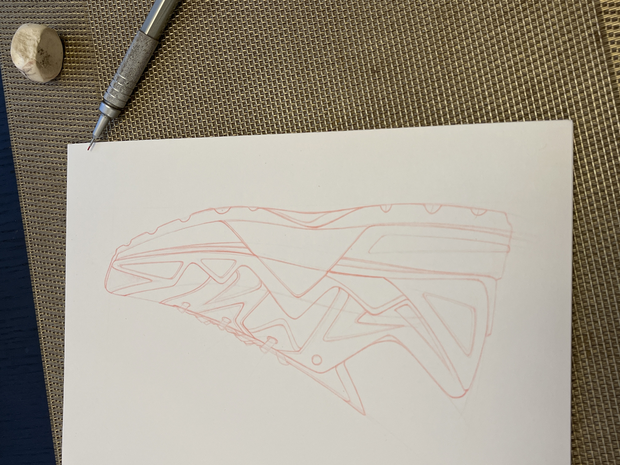 A top-down shot depicts a sketch of a shoe on a white sheet of paper. The sketch is done in red pencil. A mechanical pencil and a used eraser are positioned above the paper on a surface with a woven texture. A blue surface is partially visible on the left side of the frame. The setting is indoors. The location in the city of Krasnoyarsk, Russia, is not discernable from the provided image.