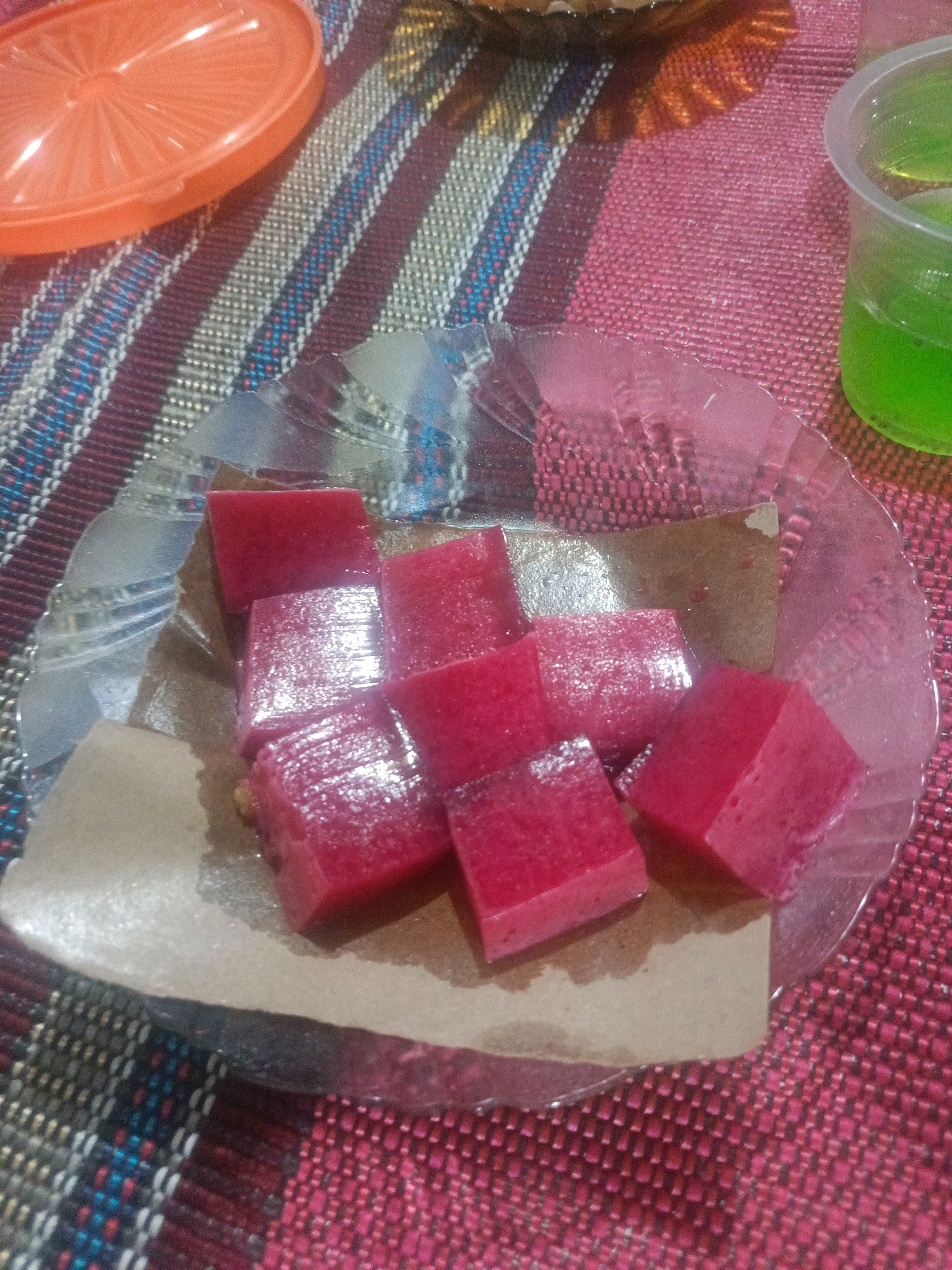 The image displays a close-up shot of a plate filled with several pieces of red jelly. The jelly is cut into cubes and arranged on a piece of brown paper, which is placed on a clear, decorative plate. The plate itself rests on a red and white striped fabric tablecloth. In the background, a portion of an orange plastic lid is visible on the left, and a clear plastic cup containing a green liquid is on the right. The lighting suggests an indoor setting, possibly a home or a small eatery in Pranti, Indonesia, given the context provided. There are no people or animals visible in the image. The focus is entirely on the food items and the surrounding surface.