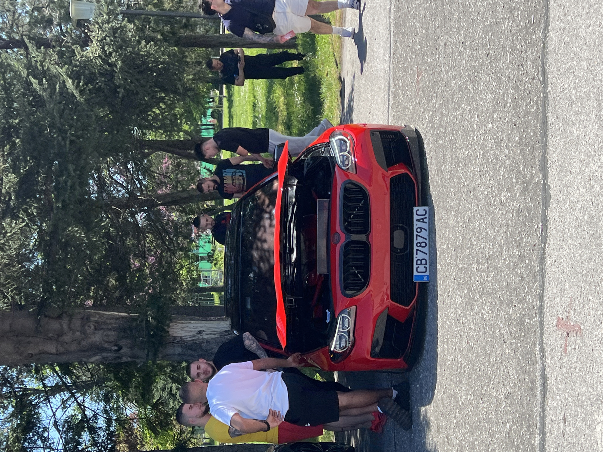 A red BMW with its hood open is parked on a gray asphalt road near trees and grass. A group of people is gathered around the car. The license plate number is CB 7879 AC. The location is likely in the city of Plovdiv, Bulgaria. The environment includes a mixture of greenery and urban elements. The people appear to be interacting with the car, possibly inspecting or working on it.