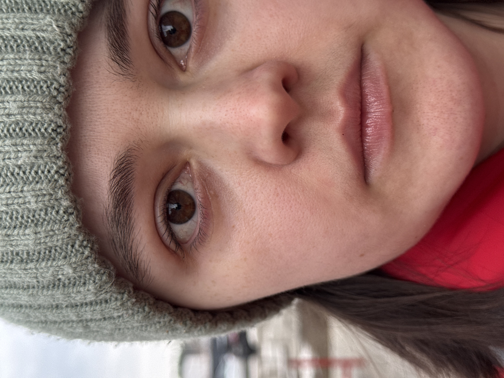 A close-up, slightly rotated image shows a person's face. The person is wearing a knitted green beanie. The person has brown eyes and fair skin with freckles. Part of a red garment is visible. The background is blurry and indistinct, and does not allow for any identification of the Izhevsk, Russia, location.