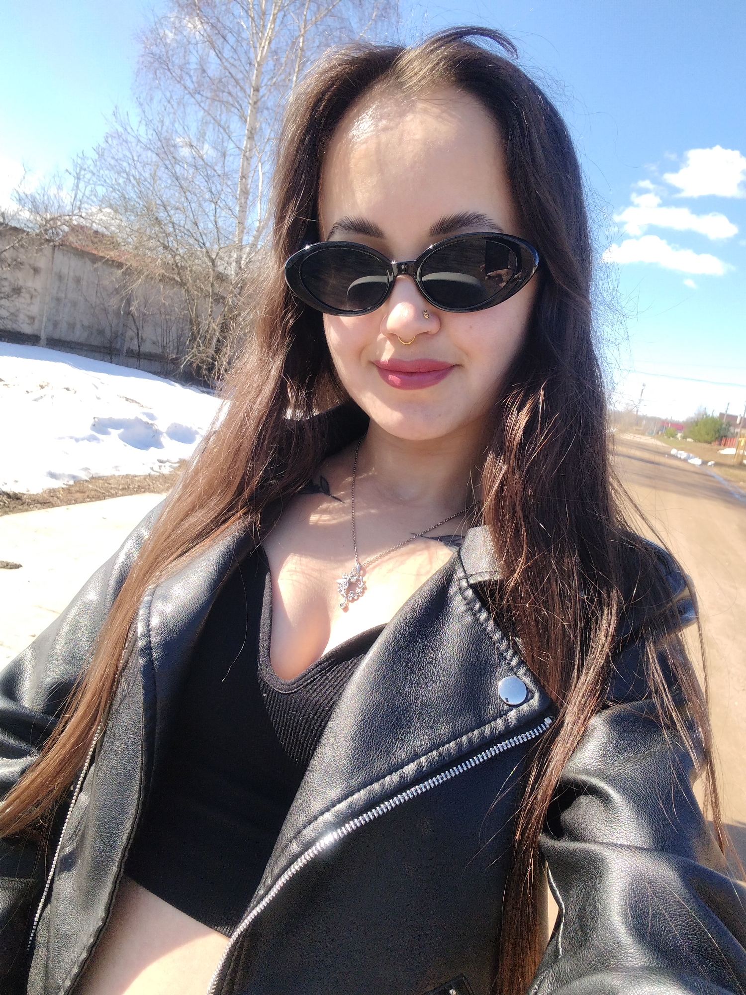 A medium-shot, eye-level selfie captures a young woman outdoors. She has long, dark hair, wears dark oval sunglasses and a black leather biker jacket over a black top. A necklace is visible. A gold nose ring is present.
The background shows a rural-urban fringe setting. A partly snow-covered embankment is visible. A partially visible concrete wall extends left. A dirt road extends off into the background toward single-story structures. A partly cloudy sky indicates daylight. The scene resembles a location on the periphery of a city or town. The location is Noviy, Russia.
