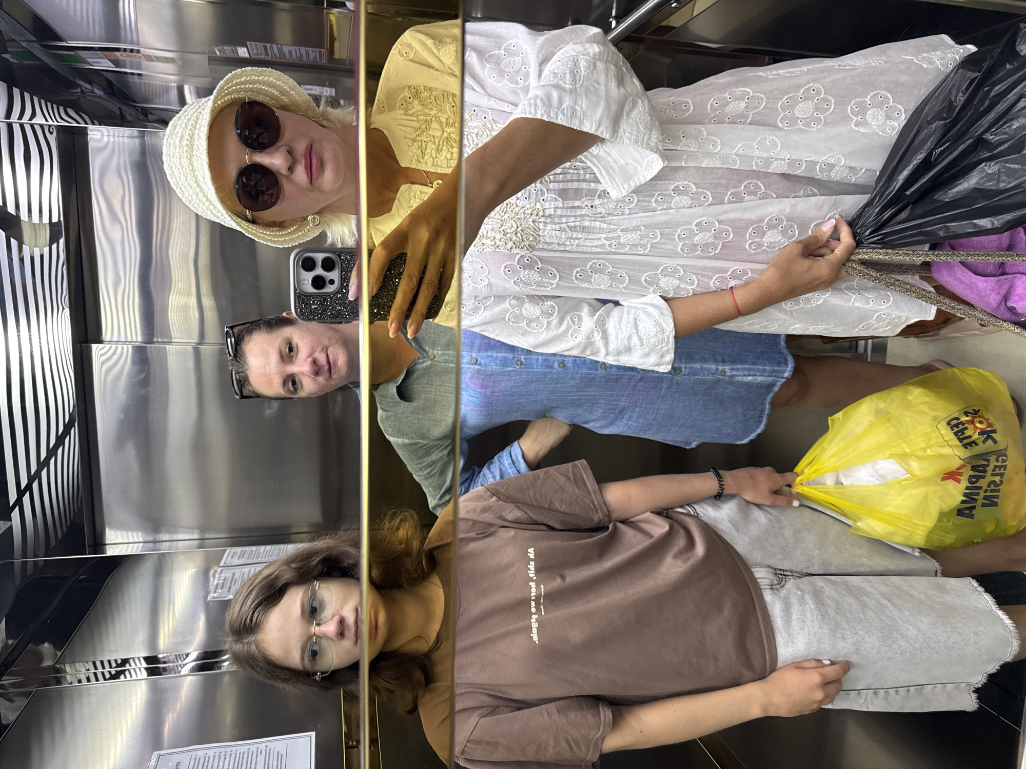 Three people are standing in an elevator. The elevator's interior consists of stainless steel walls. In the top left corner, a woman with blonde hair wearing a white hat and sunglasses is holding a smartphone, seemingly taking a selfie. Adjacent to her, another woman is reflected in the mirror, wearing sunglasses on her head. The third woman, in the bottom right corner, is wearing glasses and a brown T-shirt. One person is holding a black garbage bag and another person is holding a yellow shopping bag. The location is Unknown, Unknown.