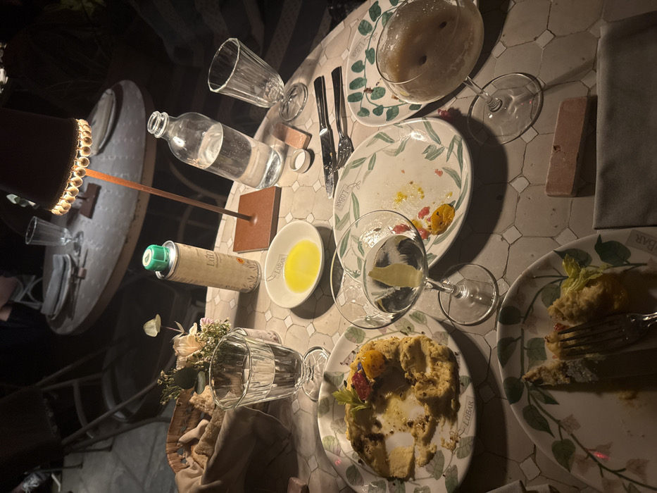 The image depicts an outdoor dining table at Lubàr restaurant in Milan, Italy, captured during the evening or night under localized artificial light from a table lamp with a dark, beaded shade. The table has a light-colored, square-tiled surface. Multiple white plates, each branded "LUBAR" with a green leafy design, show remnants of consumed food; one plate holds a largely eaten savory tart or galette with colorful toppings and a metal fork, while another contains scattered red and yellow food particles. A woven basket of bread is visible, alongside a tall can of "Lubàr" branded olive oil with a green dispenser cap and a small white bowl containing olive oil. Beverages include a clear martini-style glass with a lemon twist, a stemmed glass holding a frothy, light-brown opaque drink, a clear glass bottle of water, and two ribbed clear drinking glasses (one full of water, one empty). A set of cutlery (knife and fork), a small floral arrangement, a folded grey napkin, and a small rectangular light-colored block object are also present on the table. No individuals are visible.