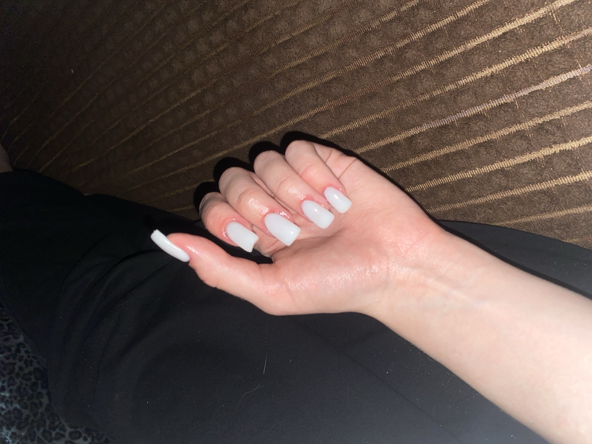 A close-up, high-angle shot shows a left hand with white, square-shaped artificial nails resting on a dark brown textured surface. The hand is positioned from the wrist upwards, with fingers slightly spread and bent. The skin appears pale with some redness around the cuticles. The background consists of a dark, possibly black, fabric on the left and the brown textured surface on the right. There is a hint of a patterned fabric in the top left corner. The lighting casts shadows from the fingers onto the brown surface. This image was taken in the city of Pesnevo, Russia.